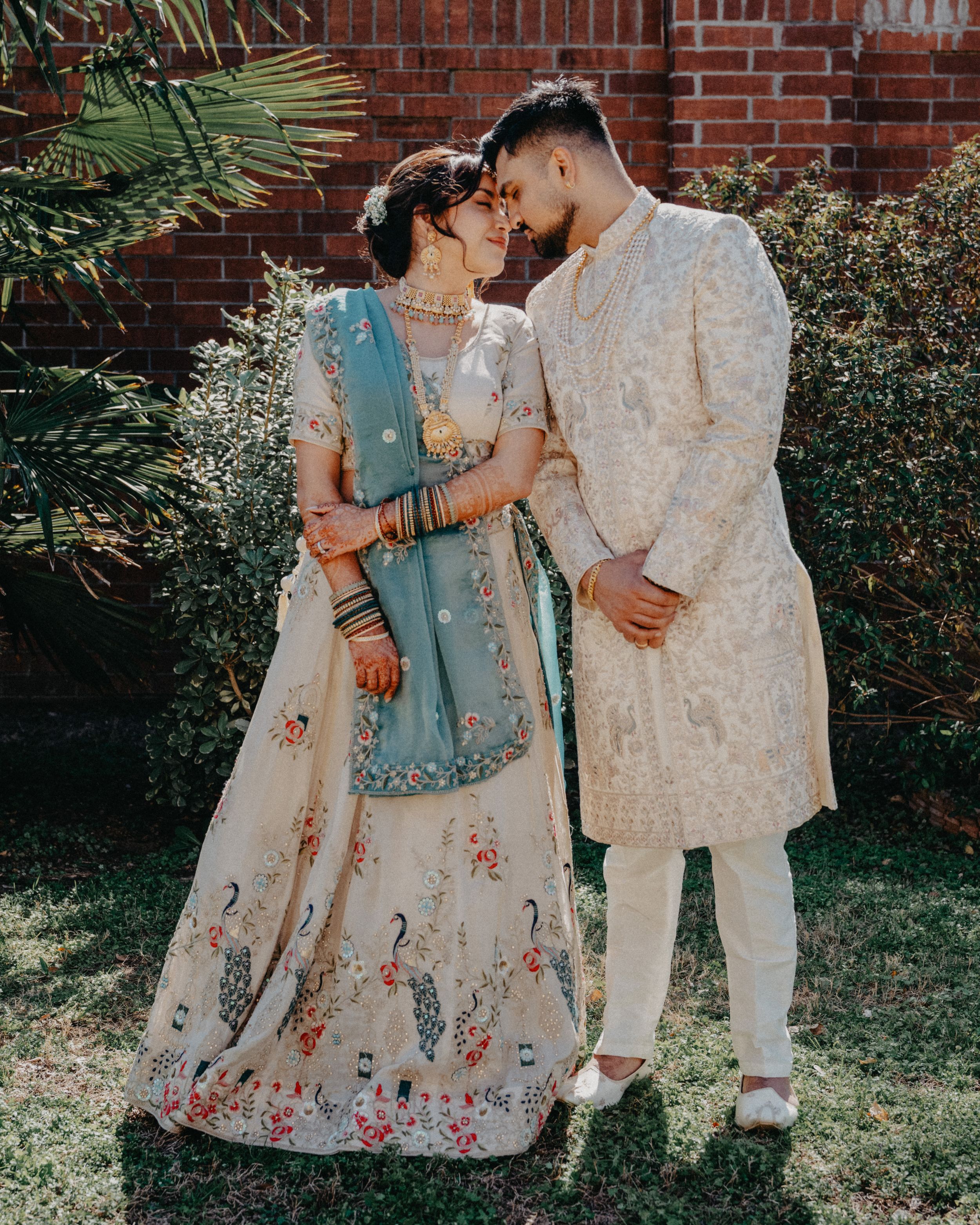 A couple in traditional South Asian wedding attire embrace against a brick wall with palm fronds in the background.