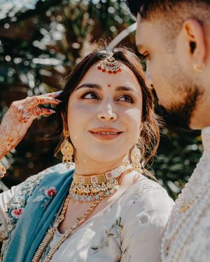 A bride wearing traditional Indian wedding jewelry and attire smiles during an outdoor ceremony.