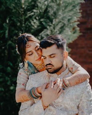 A couple in traditional South Asian wedding attire share intimate moments in a garden setting with brick wall backdrop.