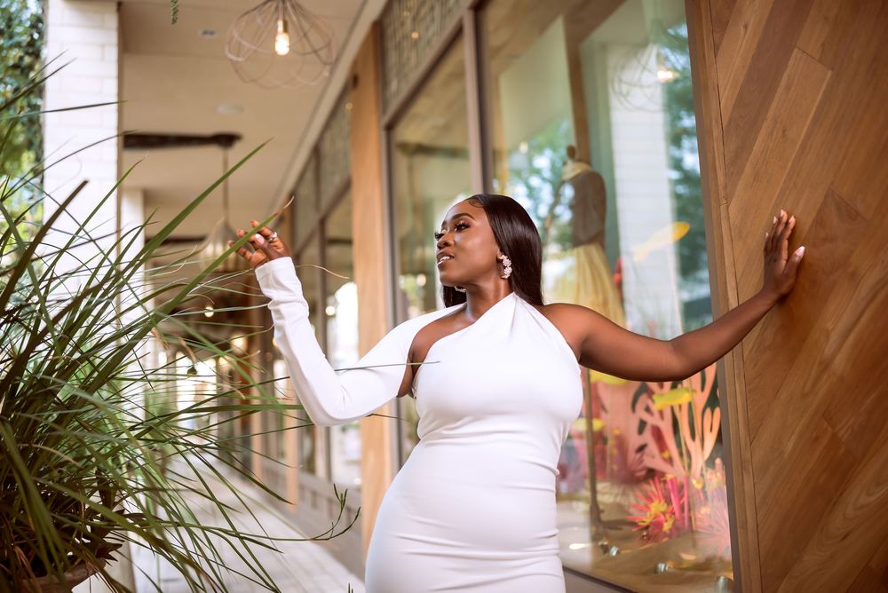 A joyful person in a white dress strikes an expressive pose on an outdoor porch with plants and windows.