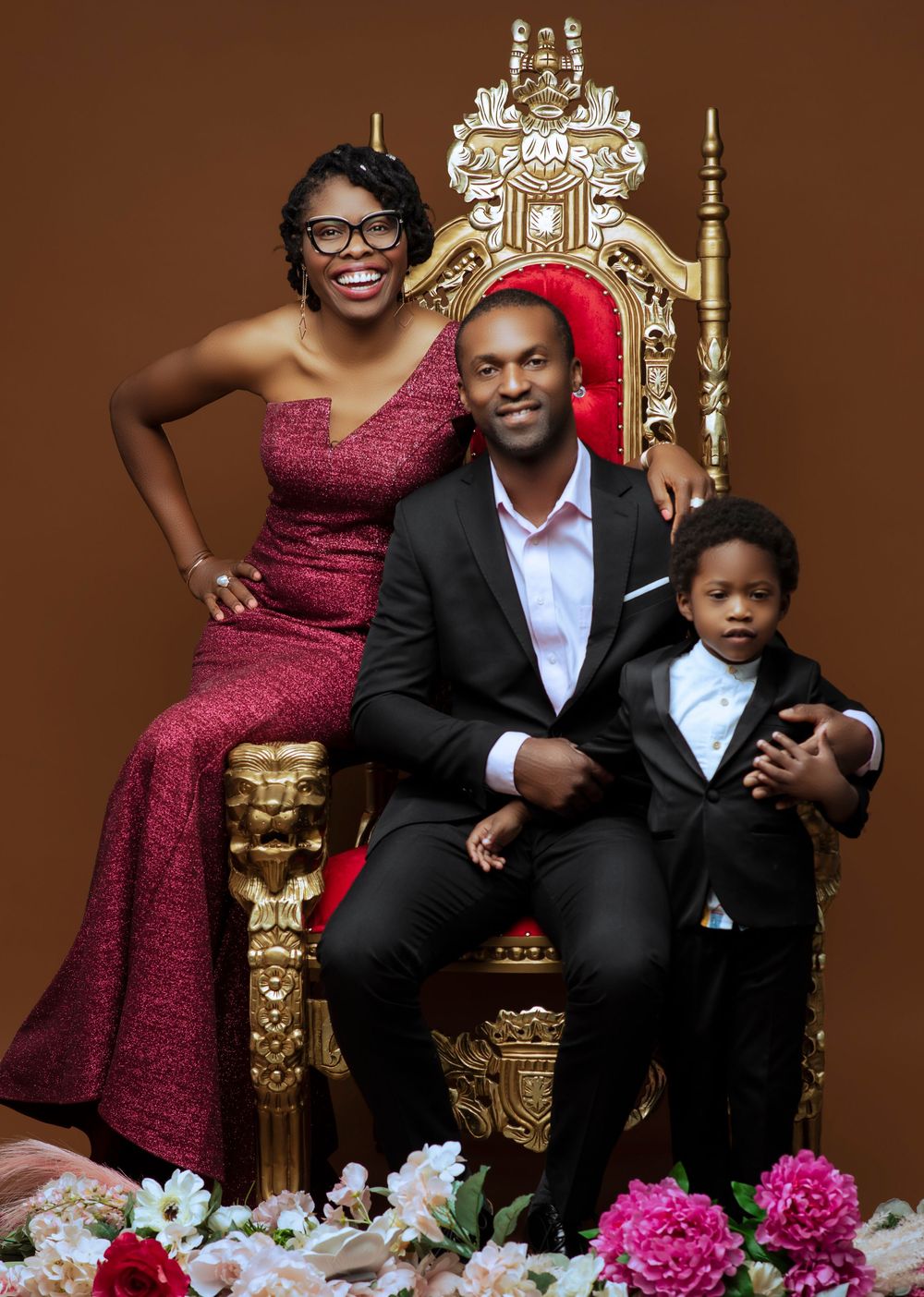 Elegant family photo session with burgundy dress and ornate gold throne against brown backdrop with pink floral decor.