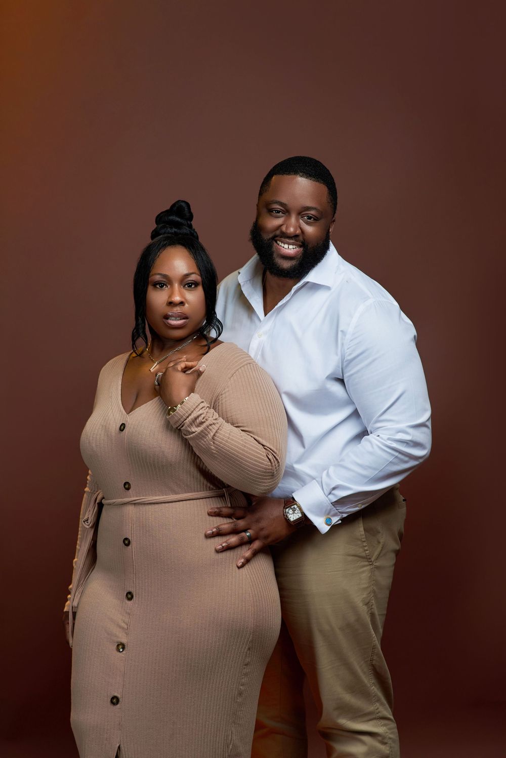 A couple poses together against a brown backdrop, wearing a beige sweater dress and white button-down shirt with khakis.