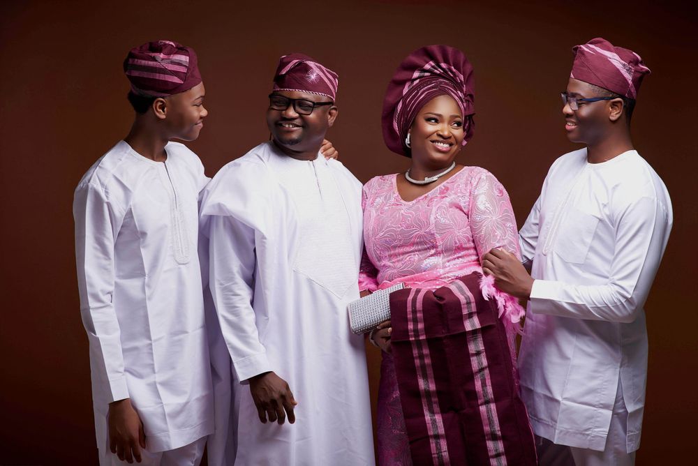 A group in traditional Nigerian attire with white agbada robes and pink gele head wraps share a joyful moment together.