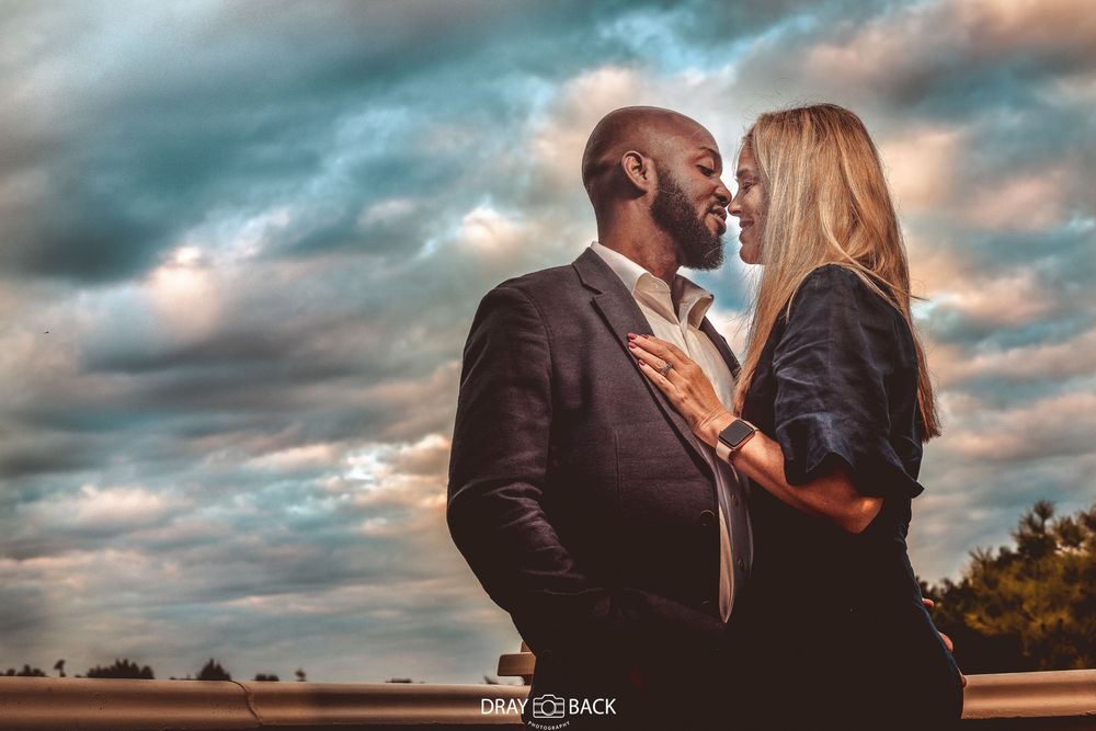 A romantic couple embraces against a dramatic sunset sky with clouds in warm golden light.