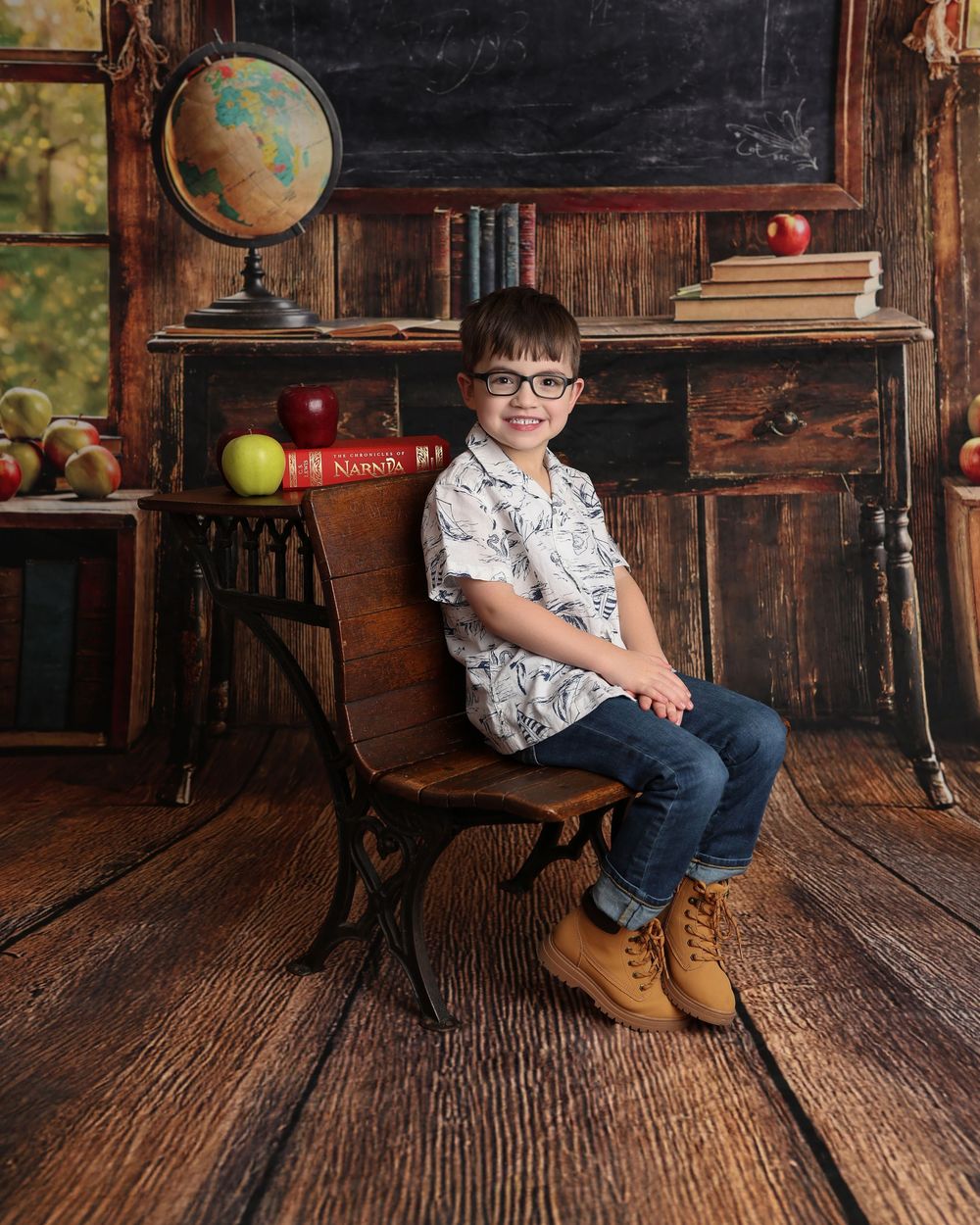 A young boy in glasses sits on a wooden school chair before a chalkboard backdrop with books, apples, and a globe in Covington, GA