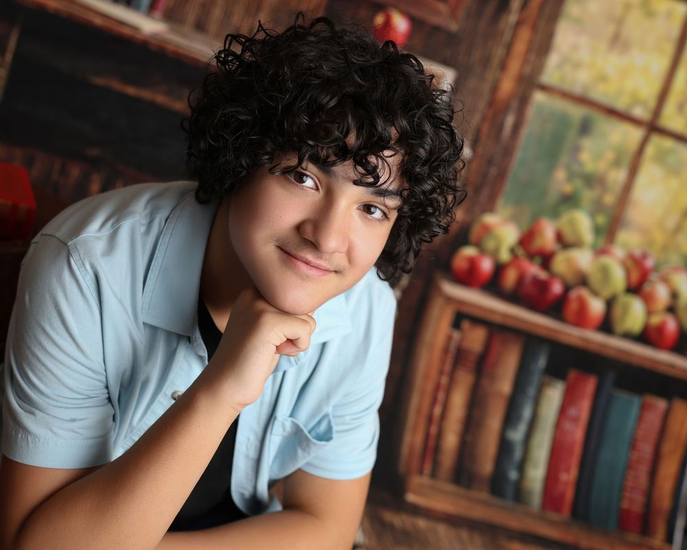 Teen boy with curly dark hair poses thoughtfully with hand on chin against a bookshelf backdrop with apples in Covington, GA