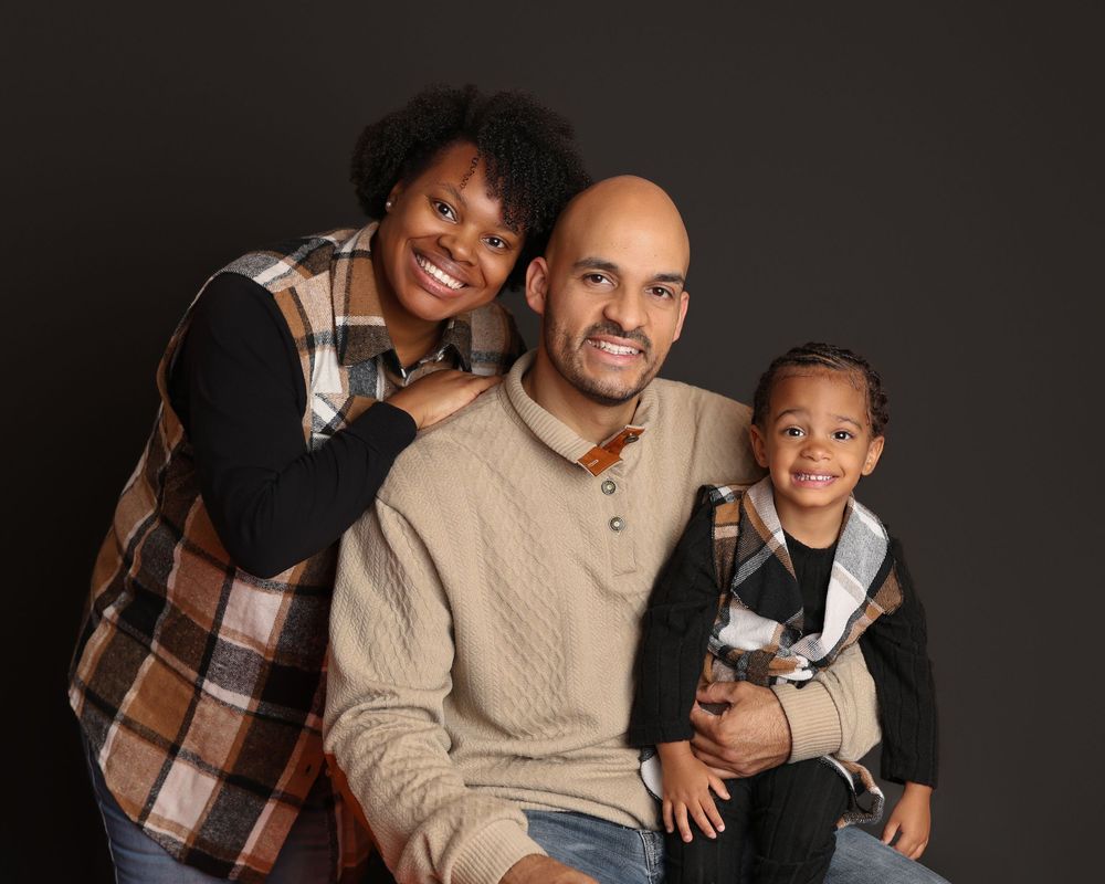 A smiling family of three poses for a studio portrait against a dark background, including parents and a young child in Covington, GA