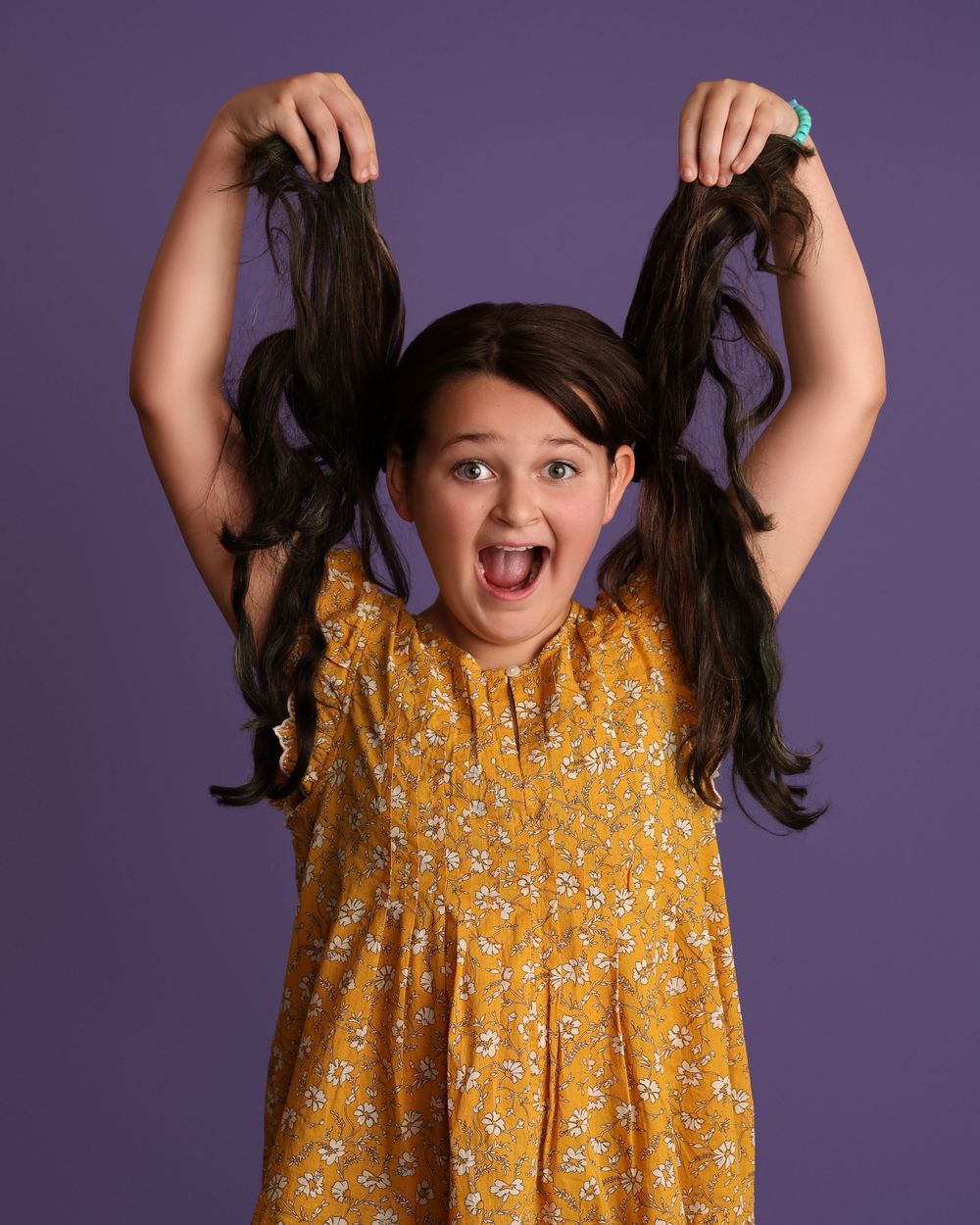A playful young girl in a yellow floral dress holds her pigtails up while making a funny face against a purple background in Covington, GA