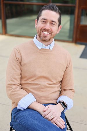 Professional headshot male sitting in chair with building as background.
