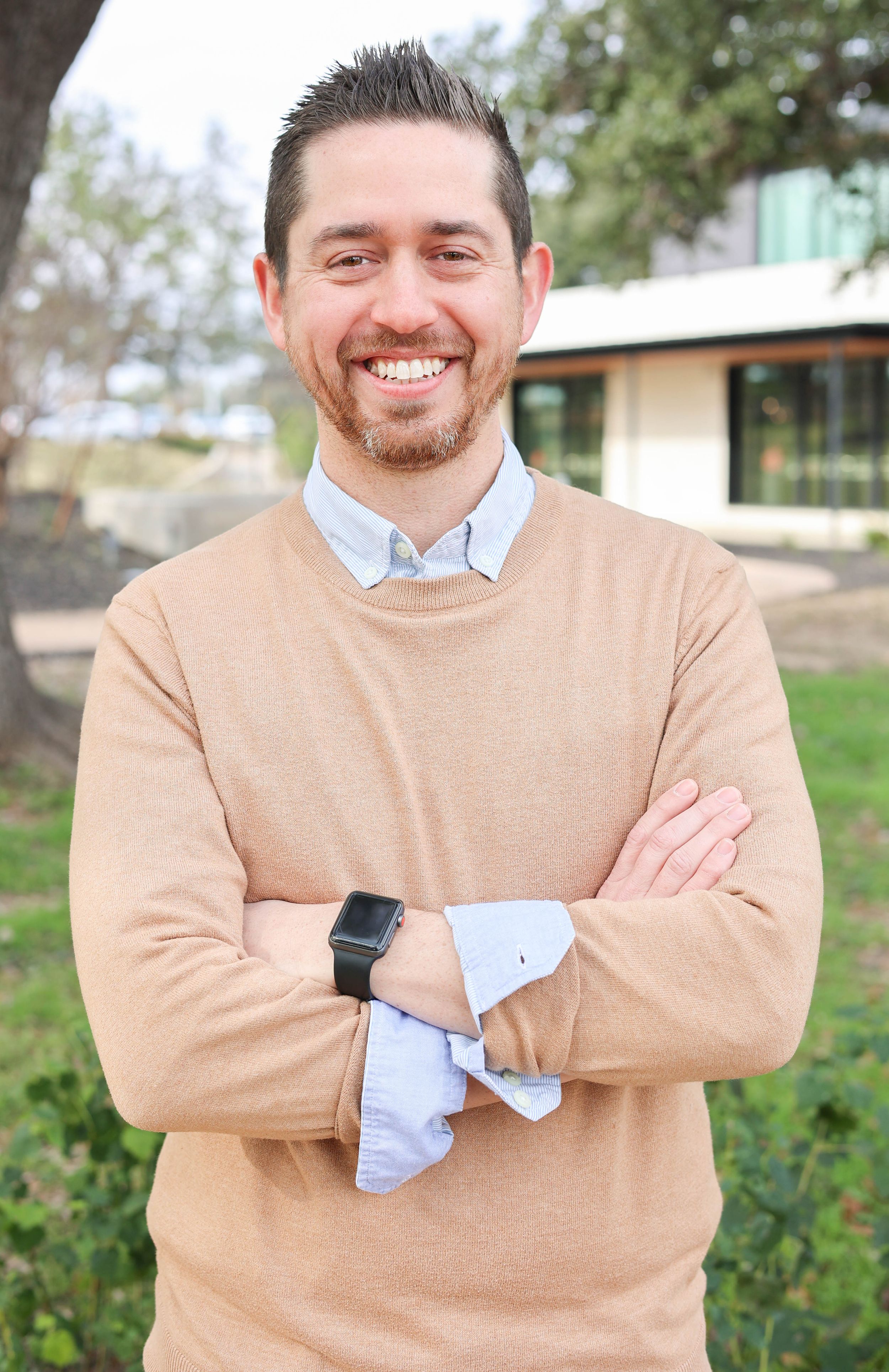 Professional headshot male  standing arms folded with building as background.