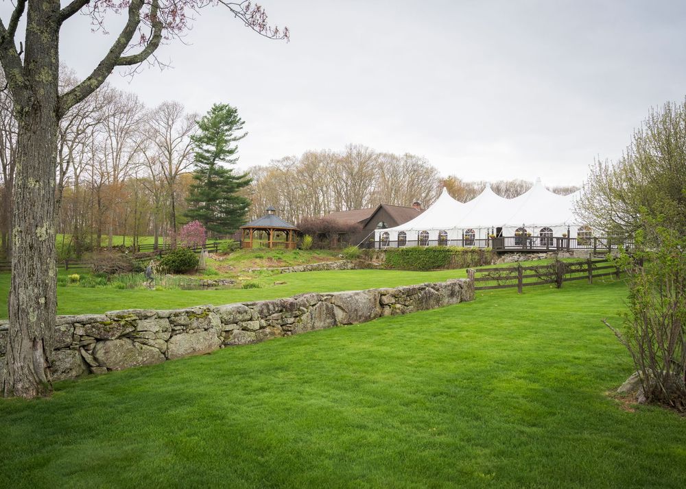 Rustic wedding venue with white tent, stone wall, wooden fence, and lush green lawn surrounded by spring trees.