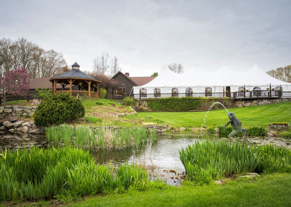 Scenic wedding venue with a pond, fountain sculpture, white tent, gazebo, and lush greenery on a cloudy spring day.