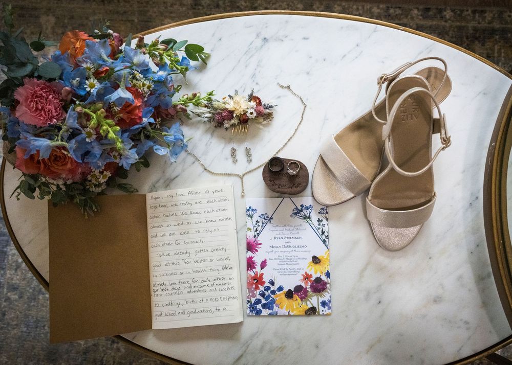 Wedding flat lay with colorful floral bouquet, invitation, handwritten vows, tan heels, and boutonniere on marble table.