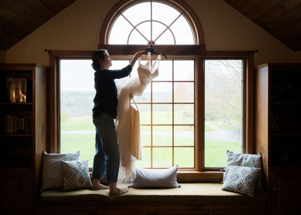 Person hanging a wedding dress on an arched window with a scenic green countryside view in a rustic venue.