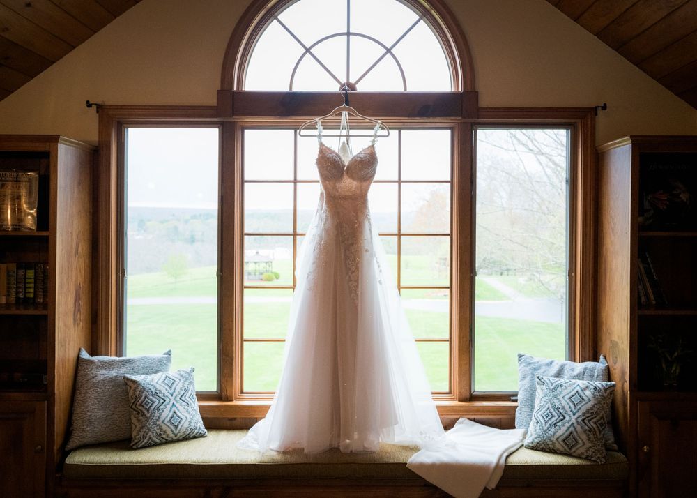 Elegant wedding gown hanging in an arched window of a rustic venue, with a lush green landscape in the background.