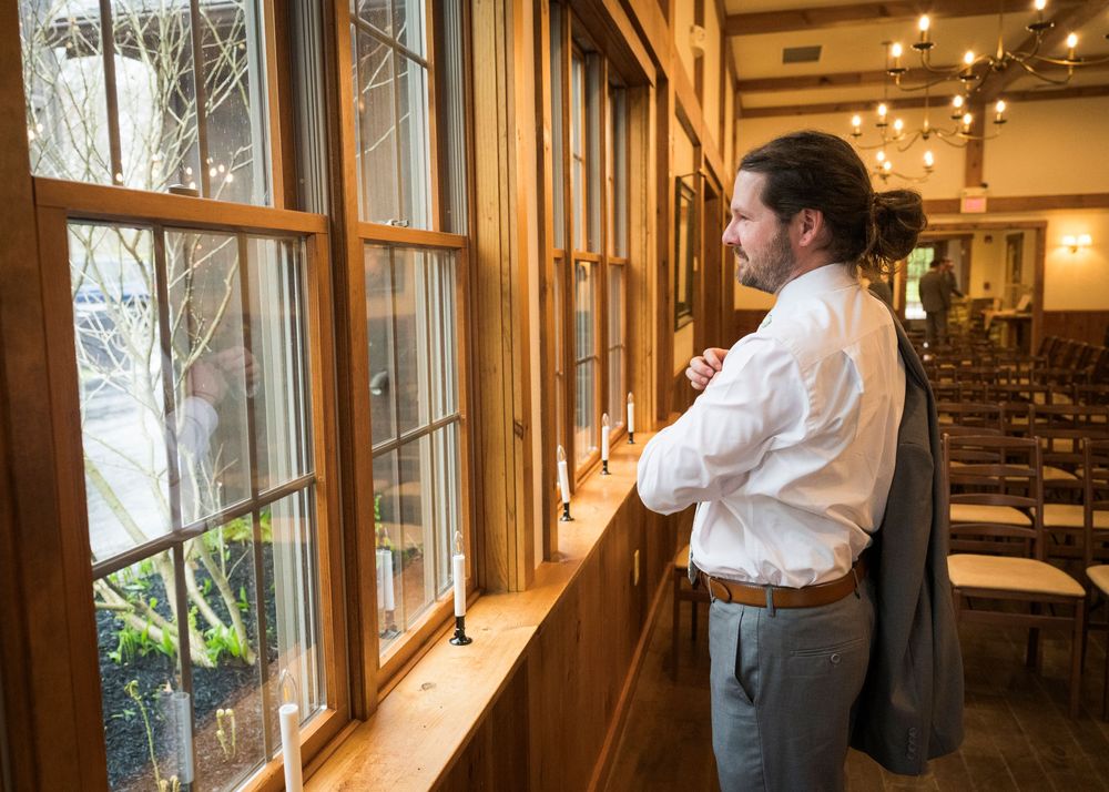 Groom in white shirt and gray suit looks out tall wooden windows of a warmly lit rustic wedding venue.
