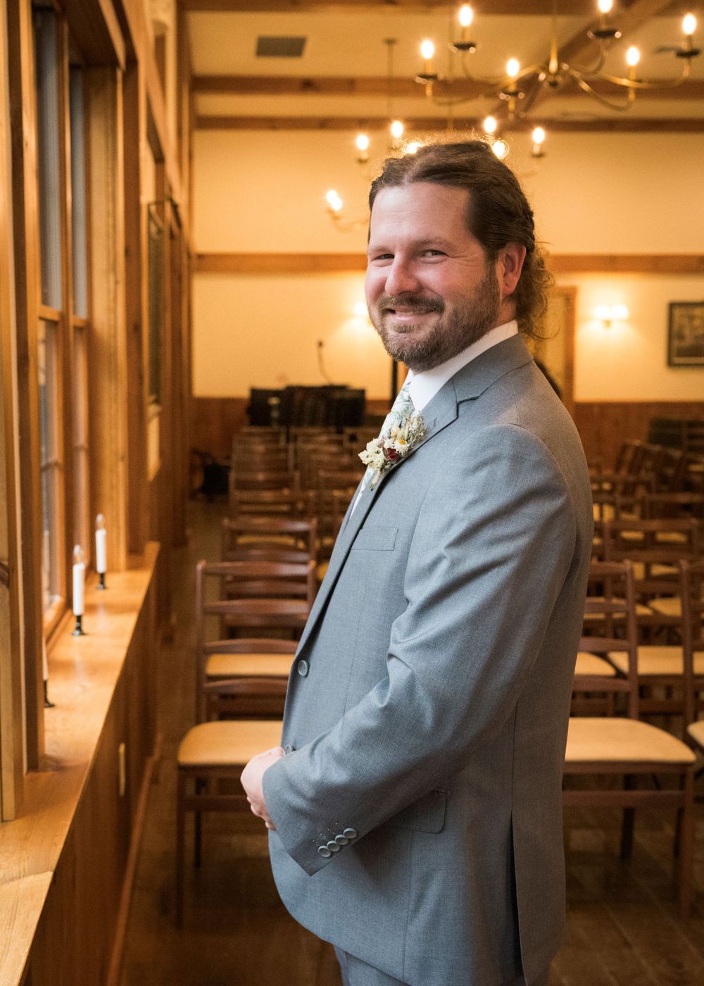 Smiling groom in gray suit with boutonniere standing in rustic wooden wedding venue with chandelier.