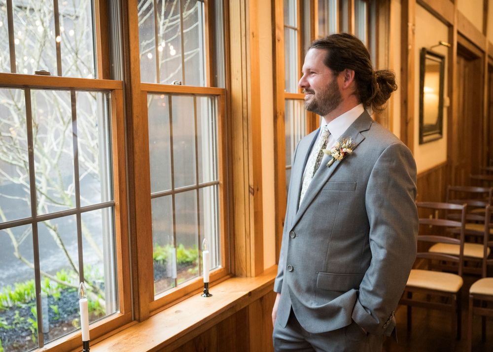 Handsome groom in a grey suit with boutonniere gazing out a wooden-framed window on his wedding day.
