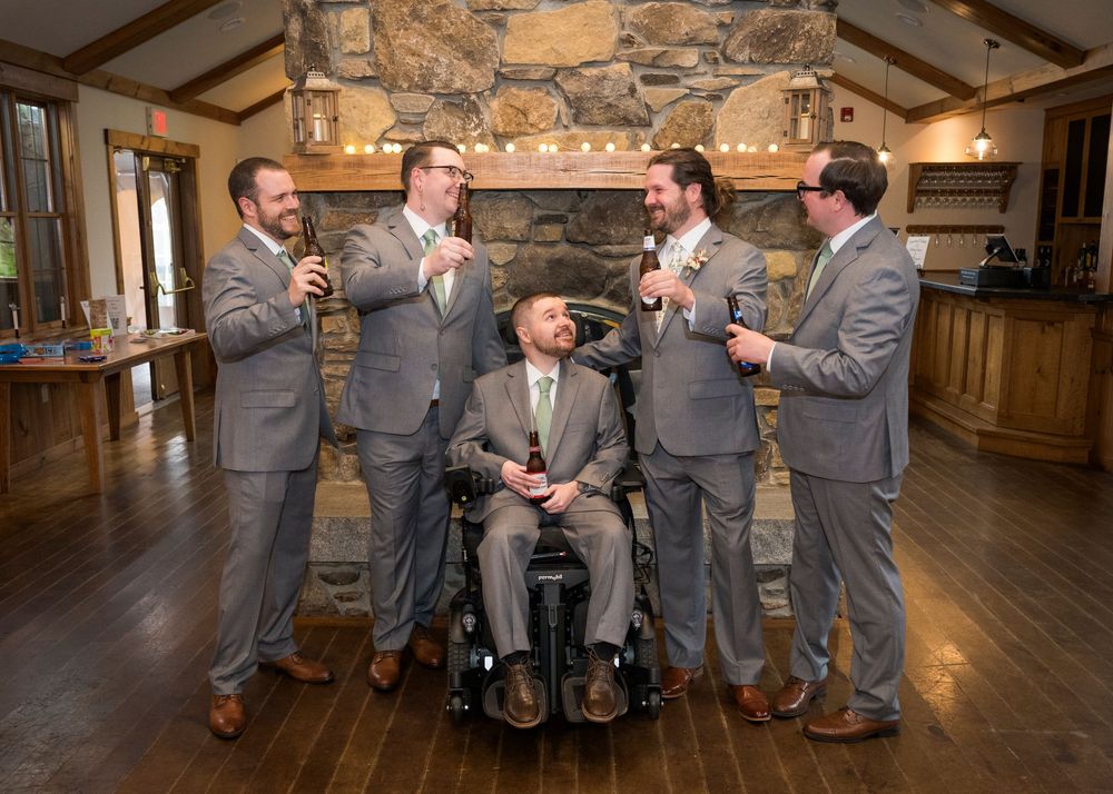 Groom and four groomsmen in grey suits celebrating with drinks near a stone fireplace at a rustic venue.