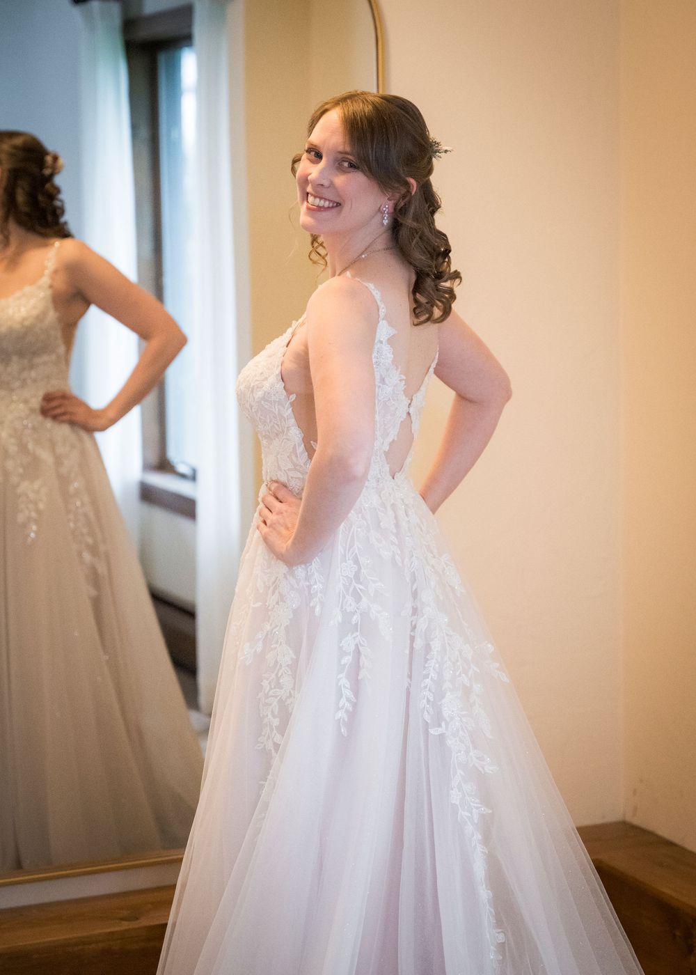 Bride in lace A-line wedding dress smiling over shoulder at mirror in bridal suite.