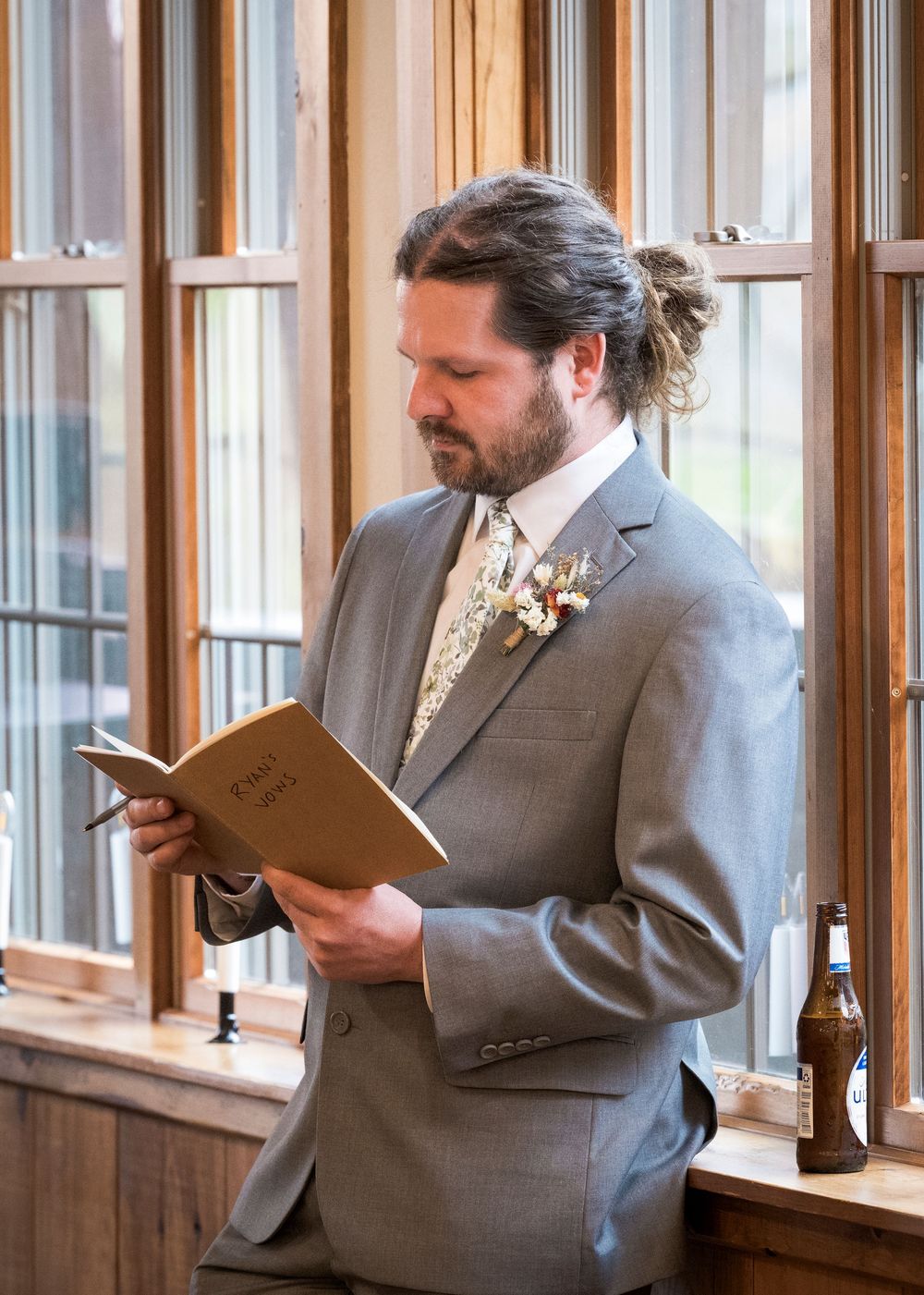 Groom in gray suit reads vows from a book at rustic wooden venue with boutonniere and beer bottle nearby.