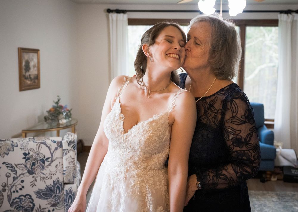 Emotional mother kissing bride on cheek during wedding morning preparations at home.