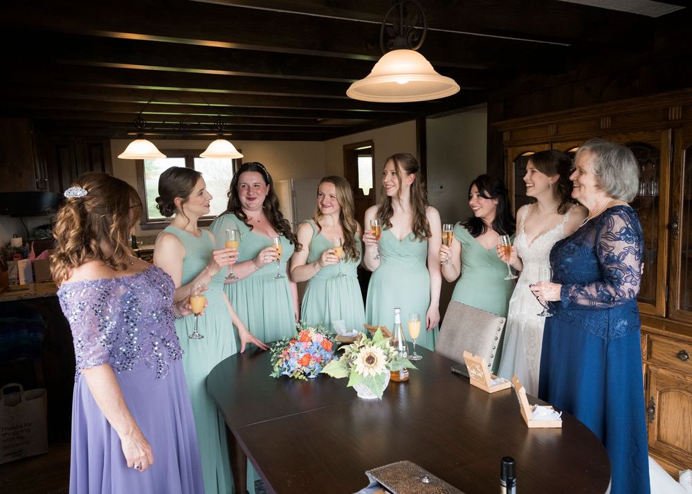 Bridesmaids in sage green dresses and guests raise champagne glasses in a toast inside a rustic wooden room.