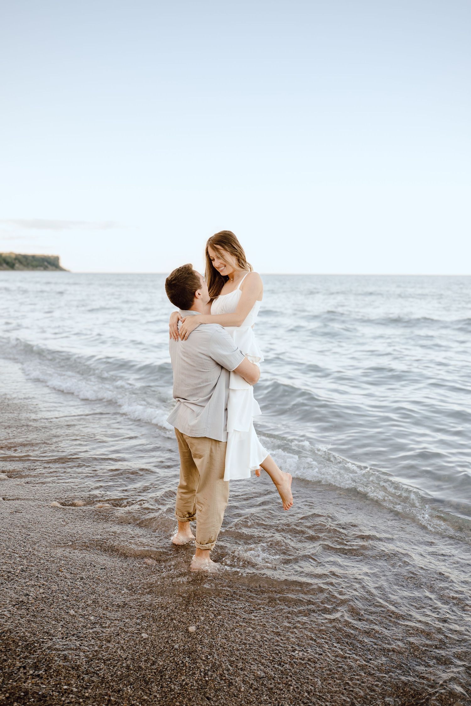 A couple embraces and spins joyfully together at the water's edge on a rocky beach at sunset.
