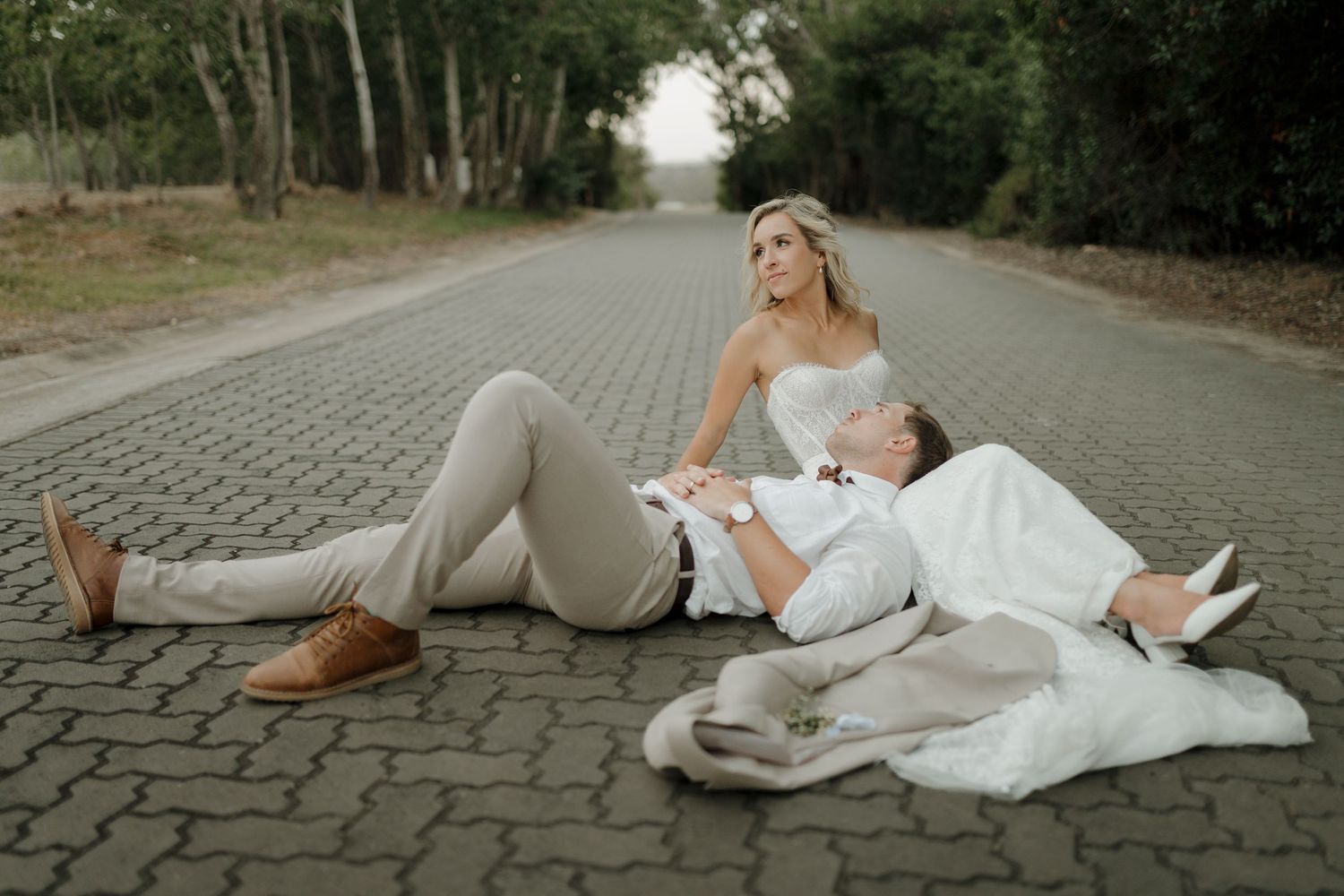 Couple in wedding attire relaxing on a cobblestone path surrounded by trees.
