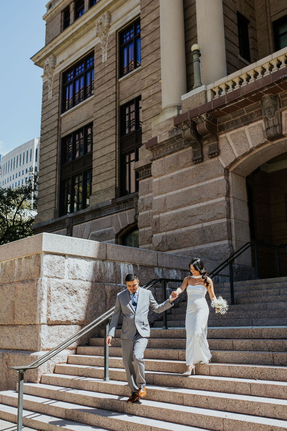 A couple descends grand stone steps outside a historic building with ornate architecture.
