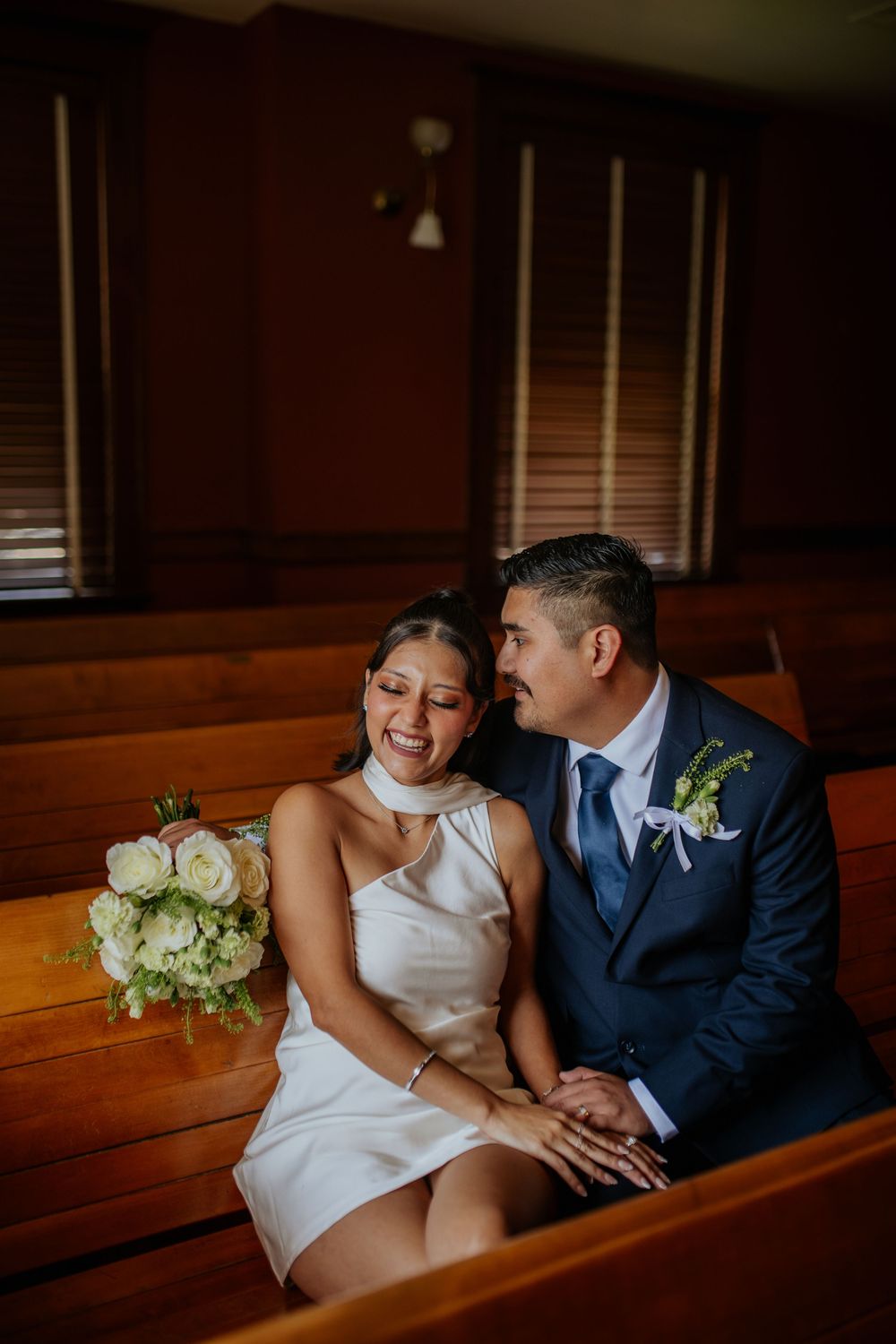 A couple in wedding attire sits intimately in a wooden church pew with soft lighting and white floral bouquet.