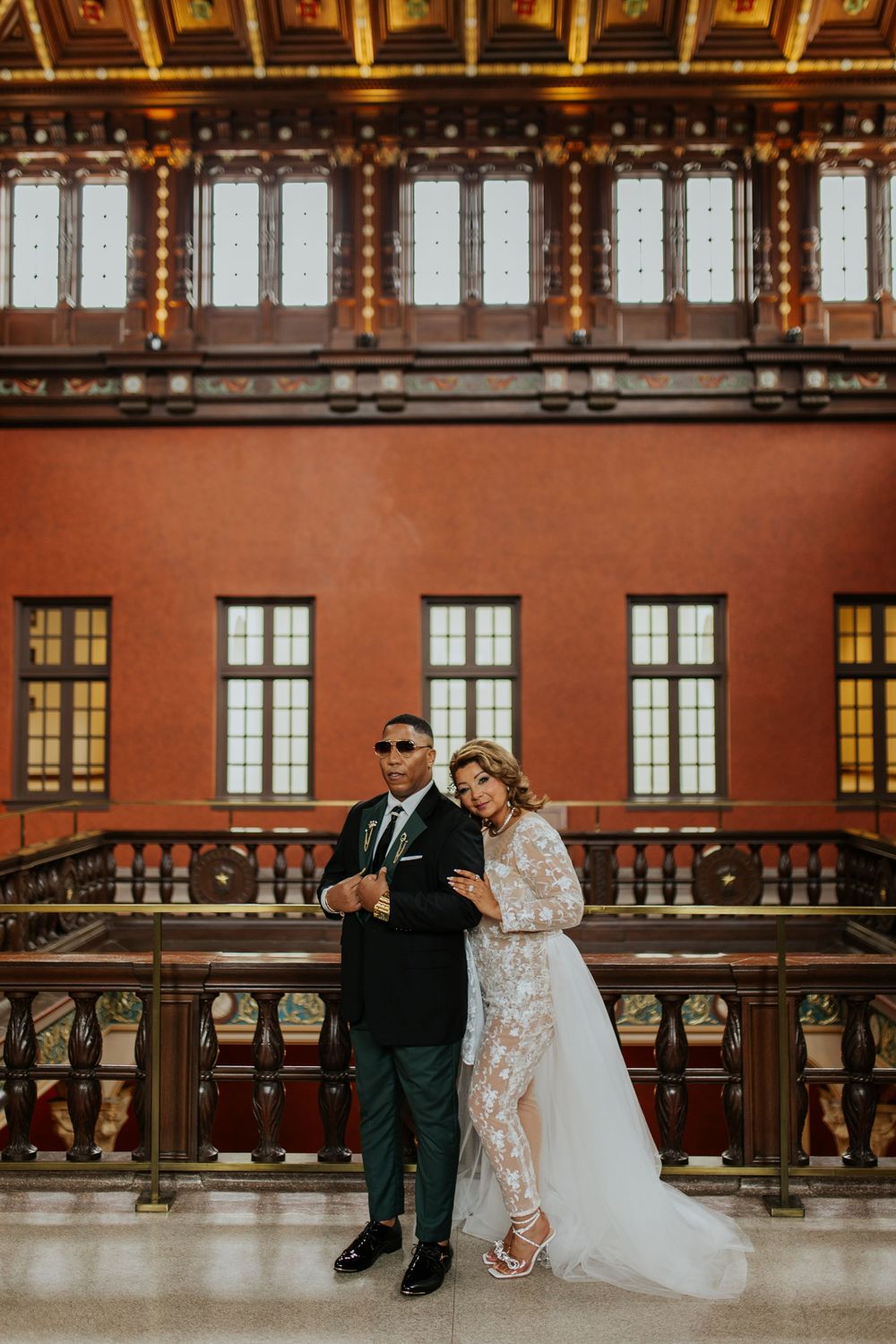 A couple poses against an ornate interior wall with vintage windows and decorative woodwork in warm lighting.