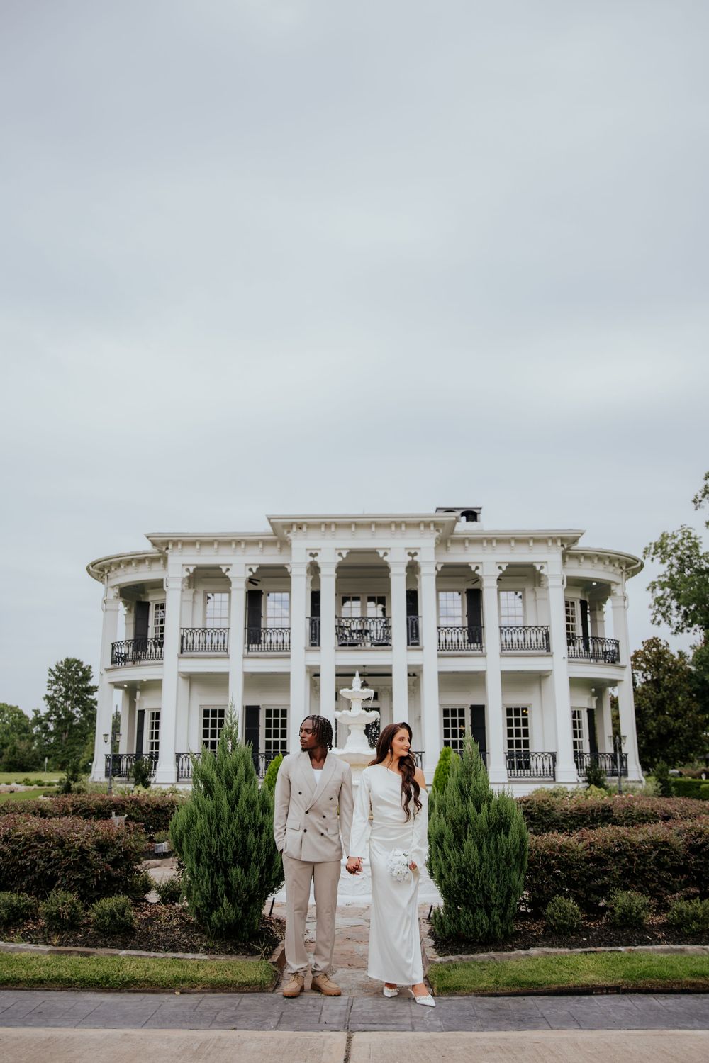 A couple walks hand in hand outside a grand white mansion with elegant columns and balconies on a cloudy day.