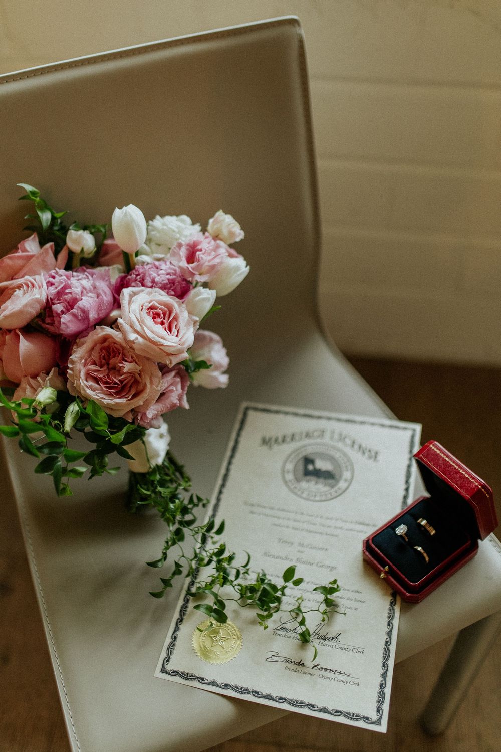 Pink roses and greenery arranged beside a certificate and ring box on a beige surface.