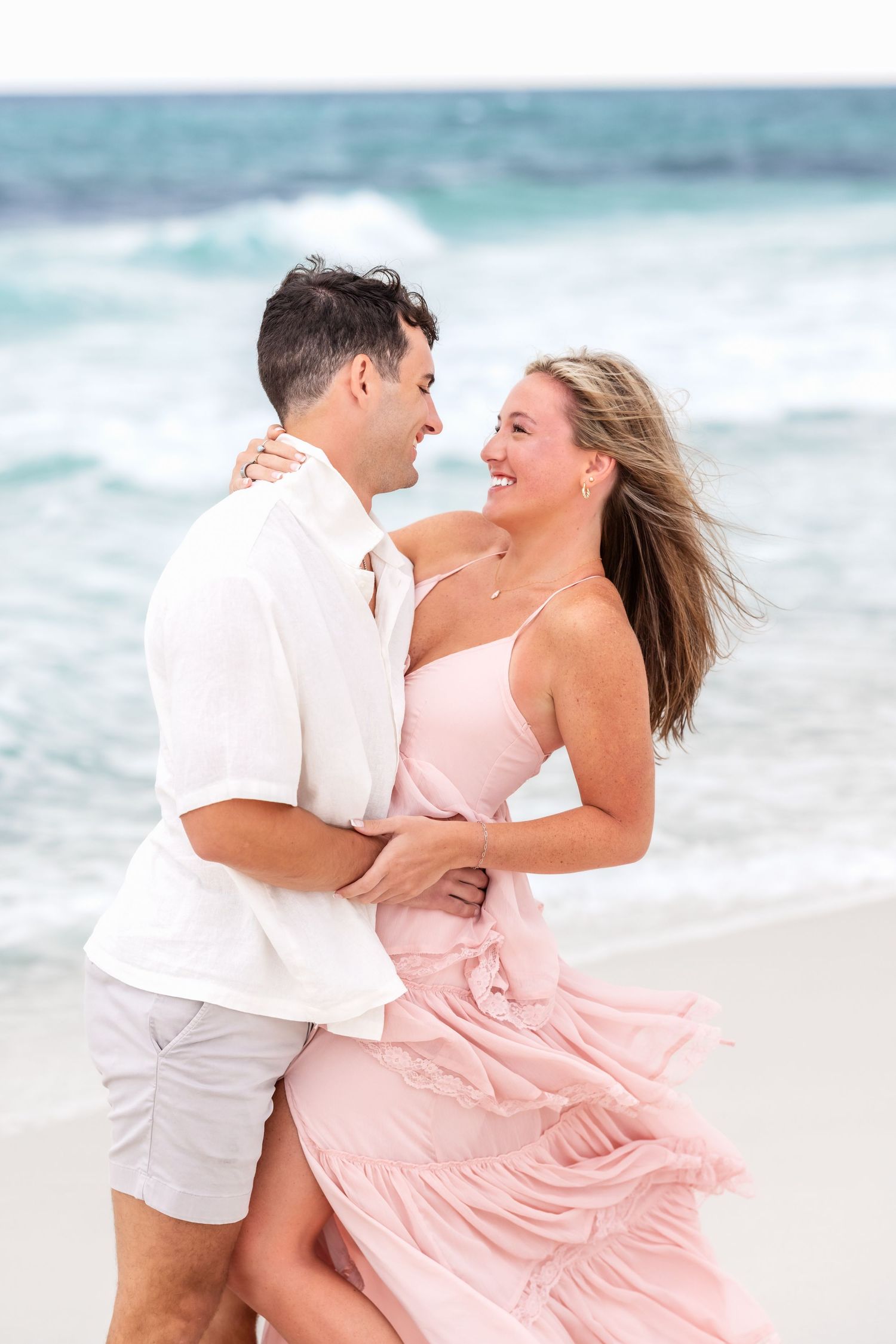 A couple in white and pink attire embrace playfully on a beach with turquoise waves in the background.