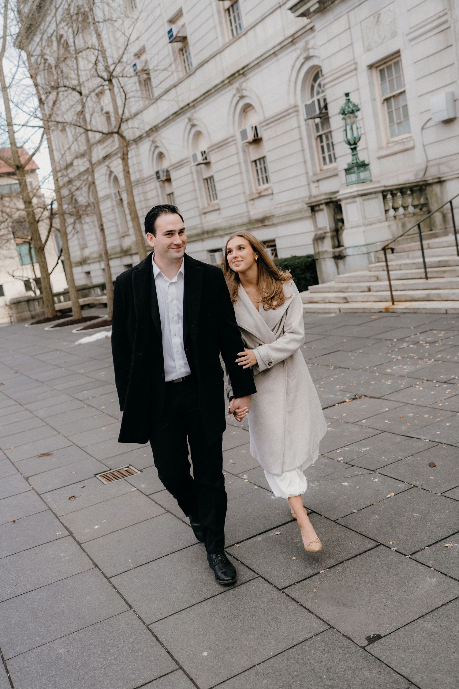 A couple in formal attire walking and laughing together on a stone sidewalk outside a grand historic building.