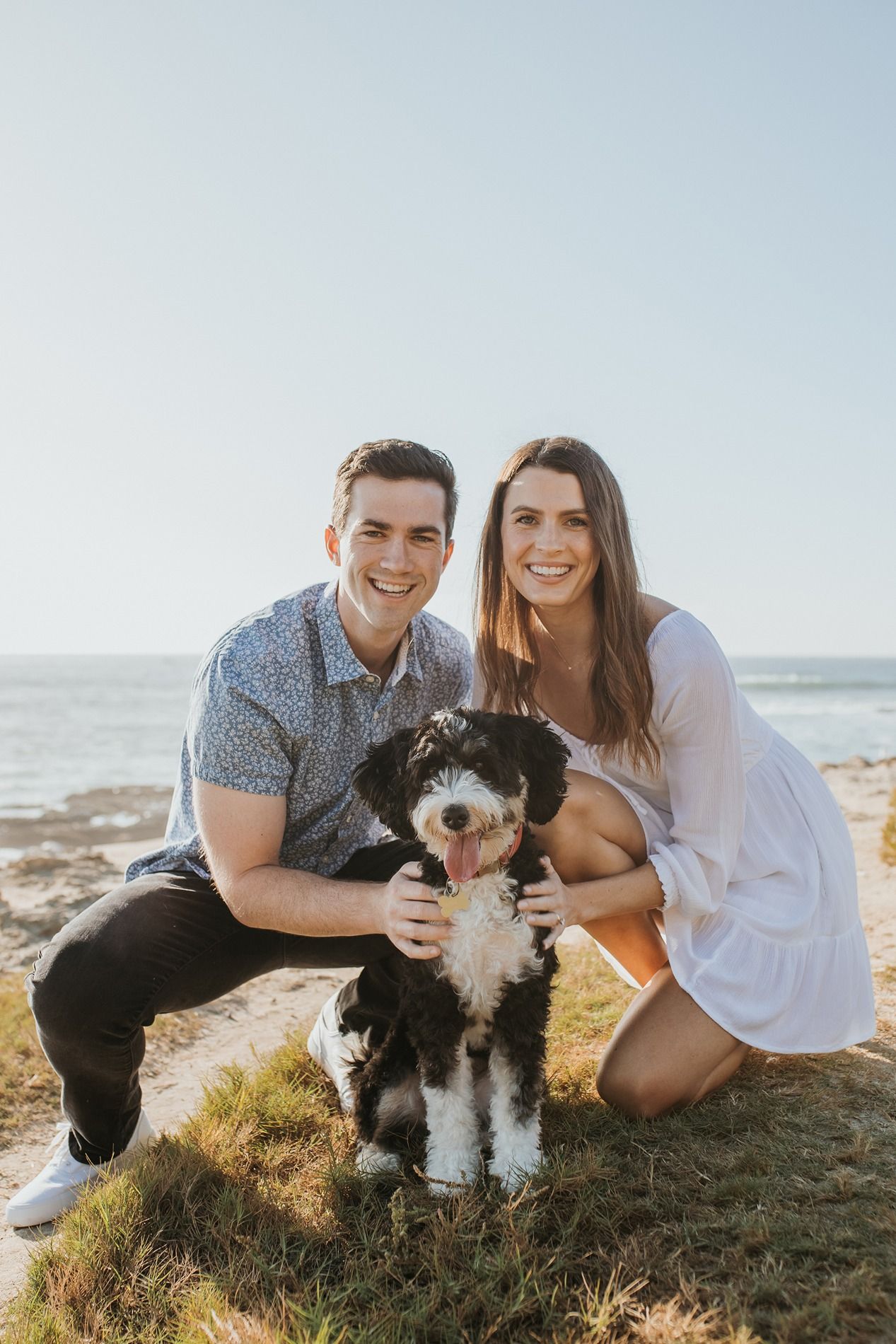 A couple poses with their black and white dog on a grassy cliff overlooking the ocean at sunset at Windansea Beach.