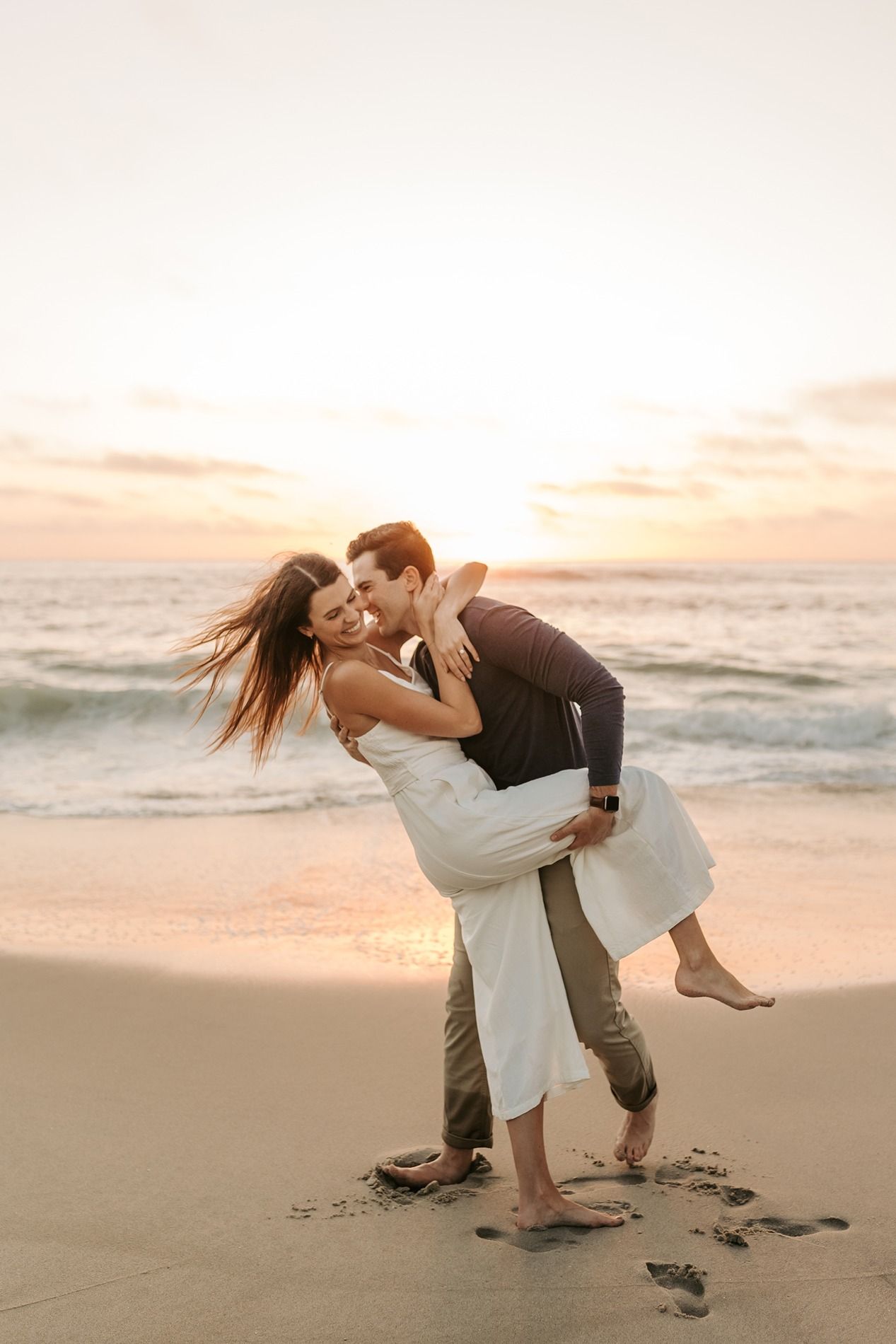 A romantic couple embraces and dances on a sandy beach at sunset with ocean waves in the background at Windansea Beach..