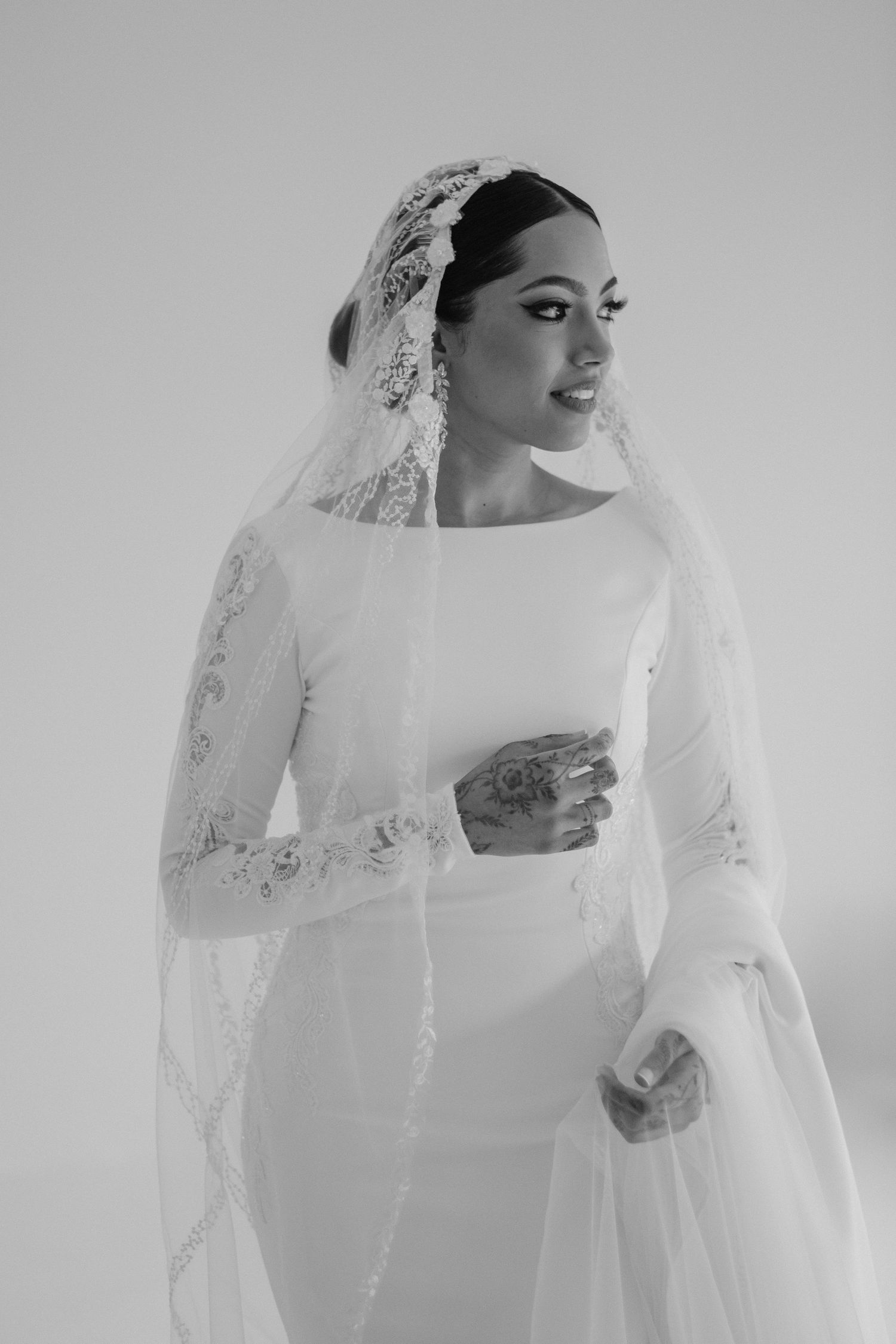 Elegant bride in long-sleeve white gown with ornate lace veil in black and white portrait.