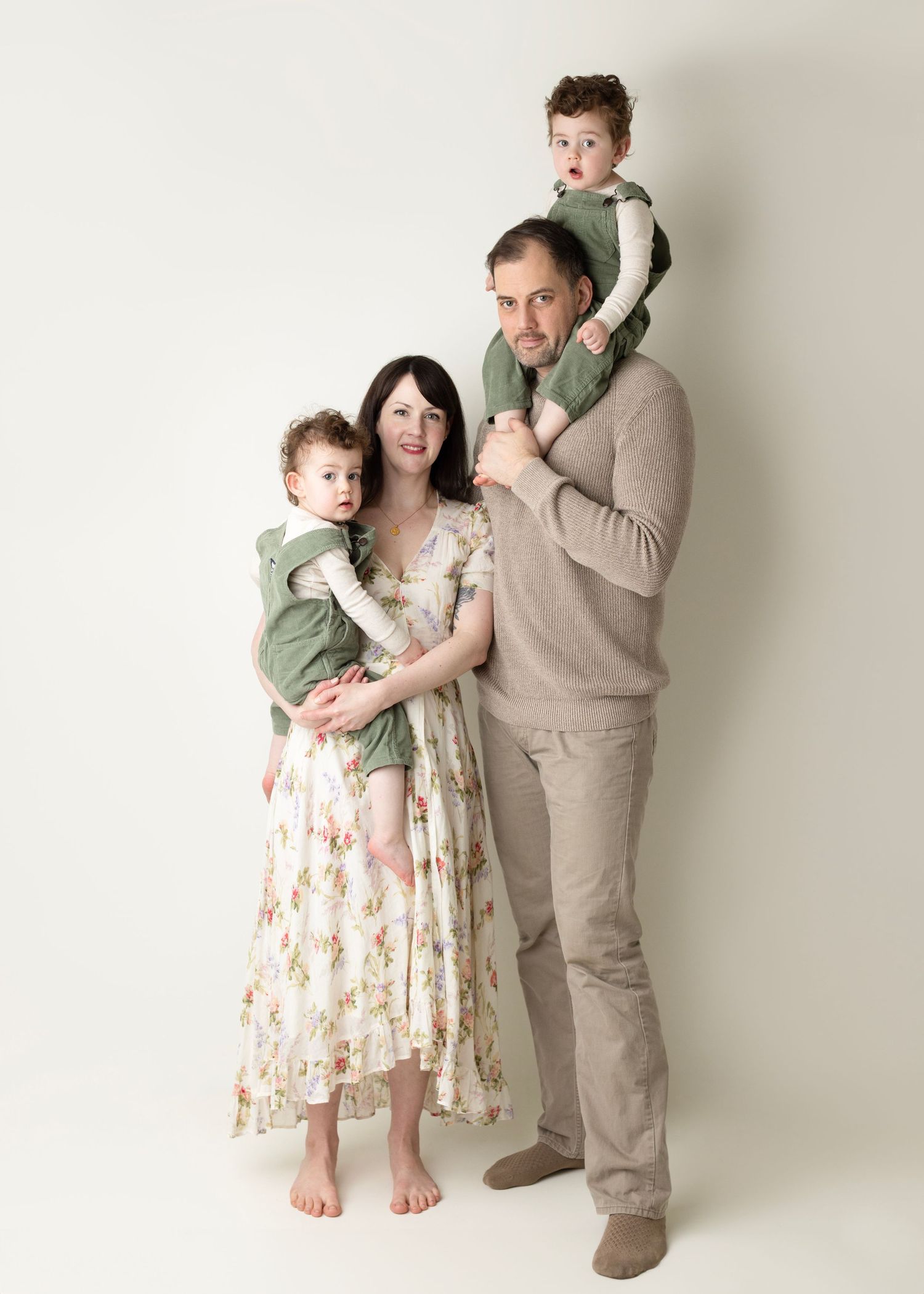 A family of four poses together against a white backdrop wearing coordinated neutral and sage green outfits.