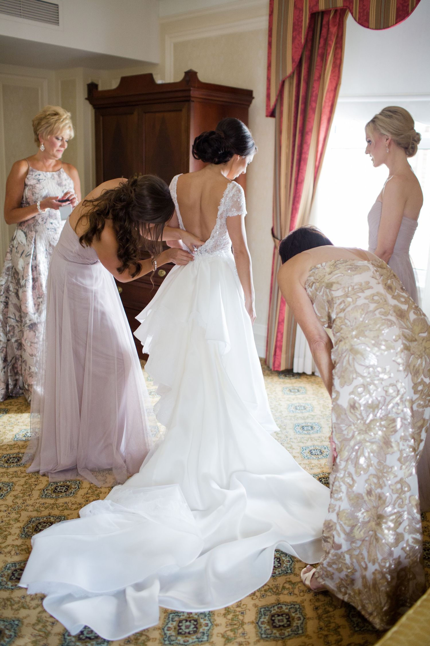 A group of bridesmaids in flowing pink and white dresses help the bride prepare in an elegant hotel room.