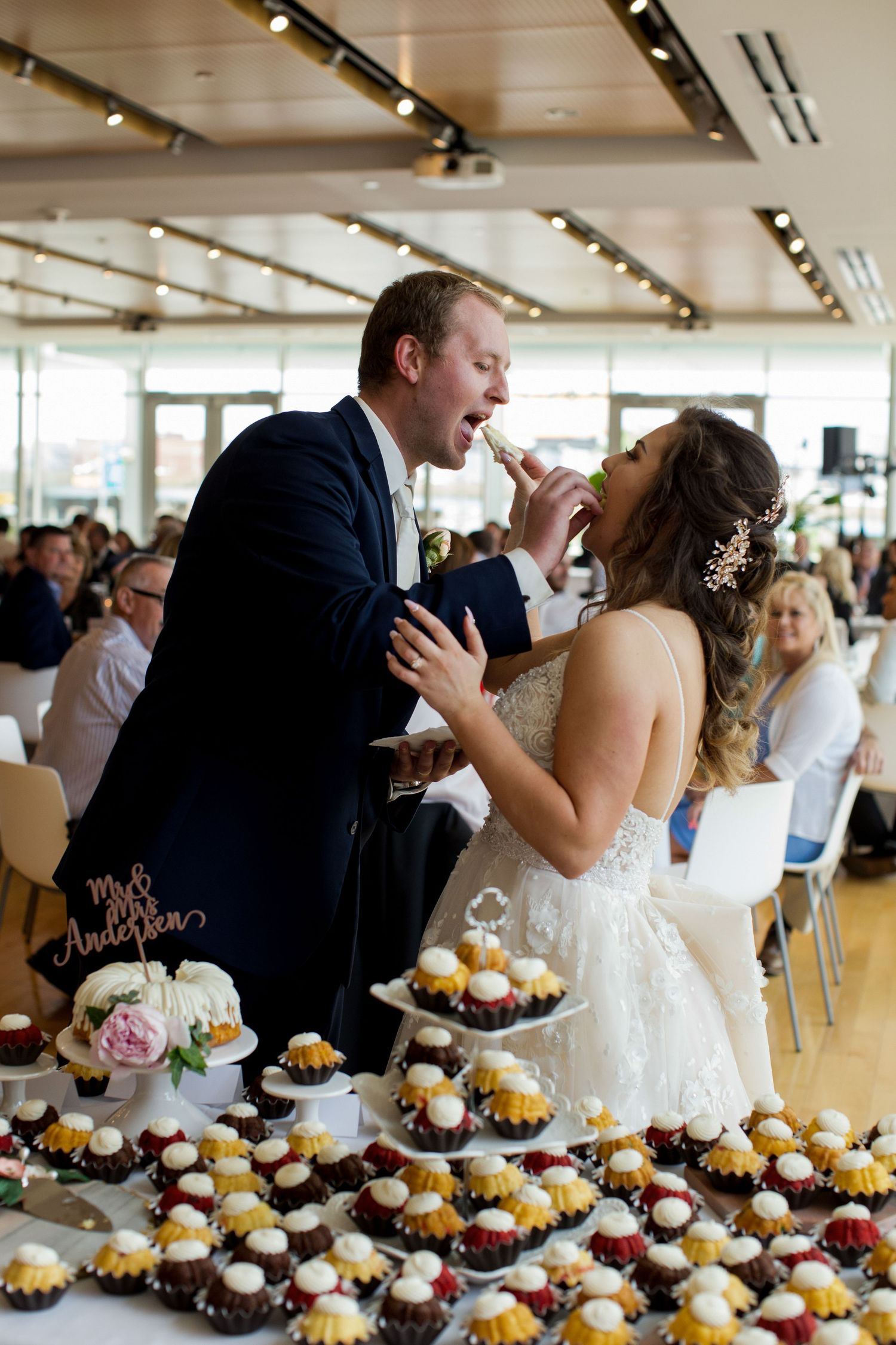 A couple shares a playful moment while cutting their wedding cake, surrounded by cupcakes at their reception.