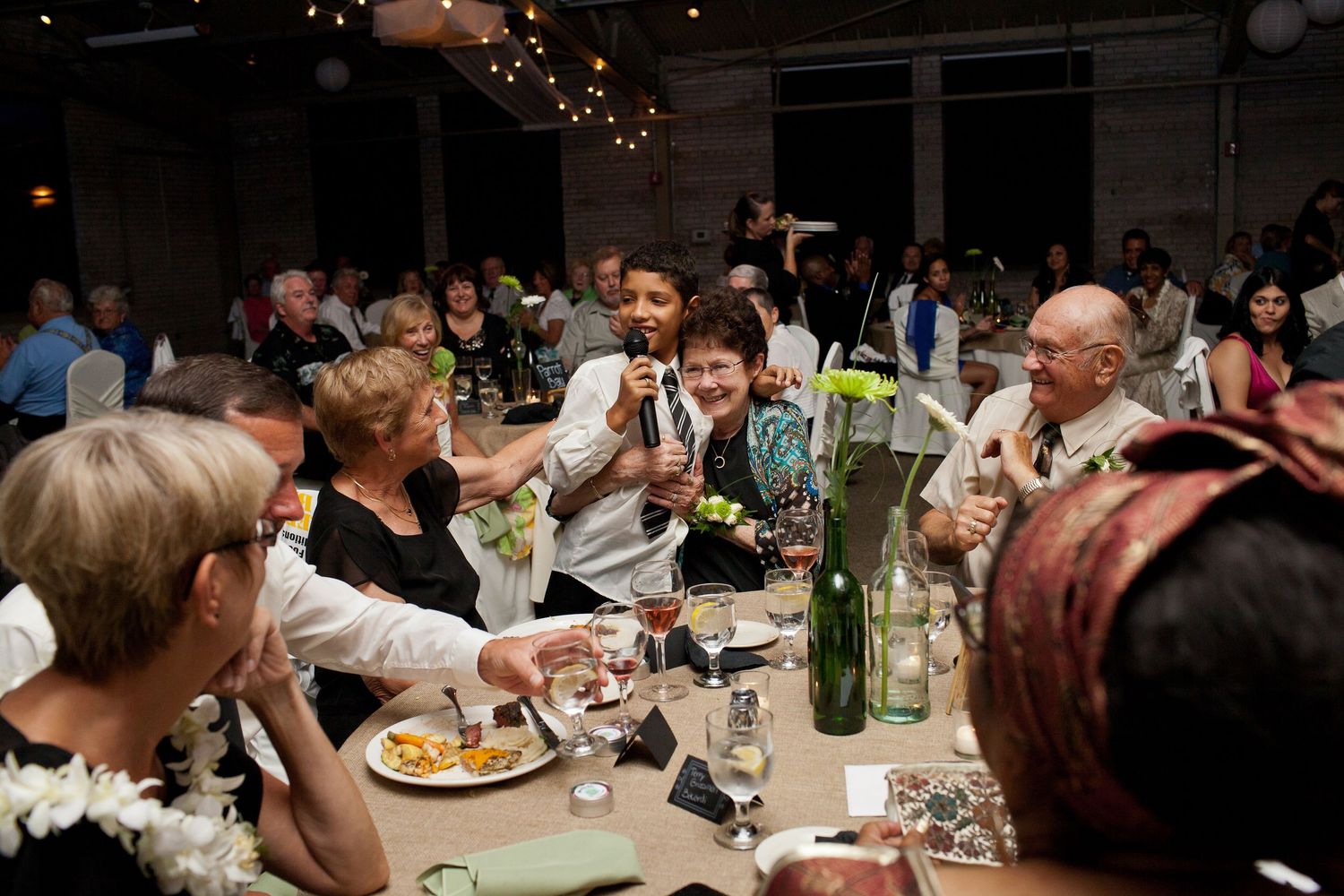 Wedding guests enjoy dinner and conversation at a reception with string lights overhead in an indoor venue.