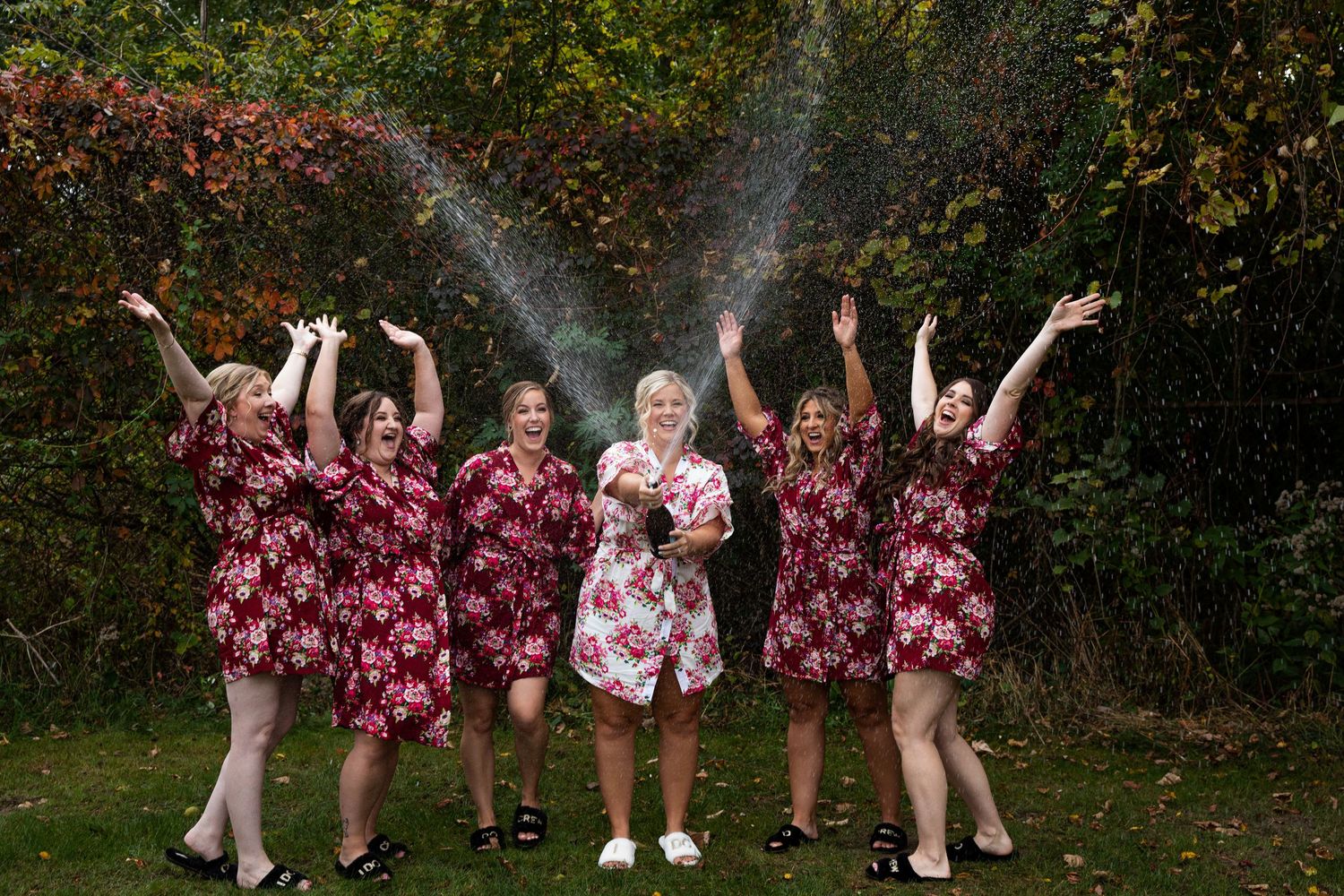 A group celebrates outdoors wearing matching burgundy floral dresses while water sprays in the air on an autumn day.