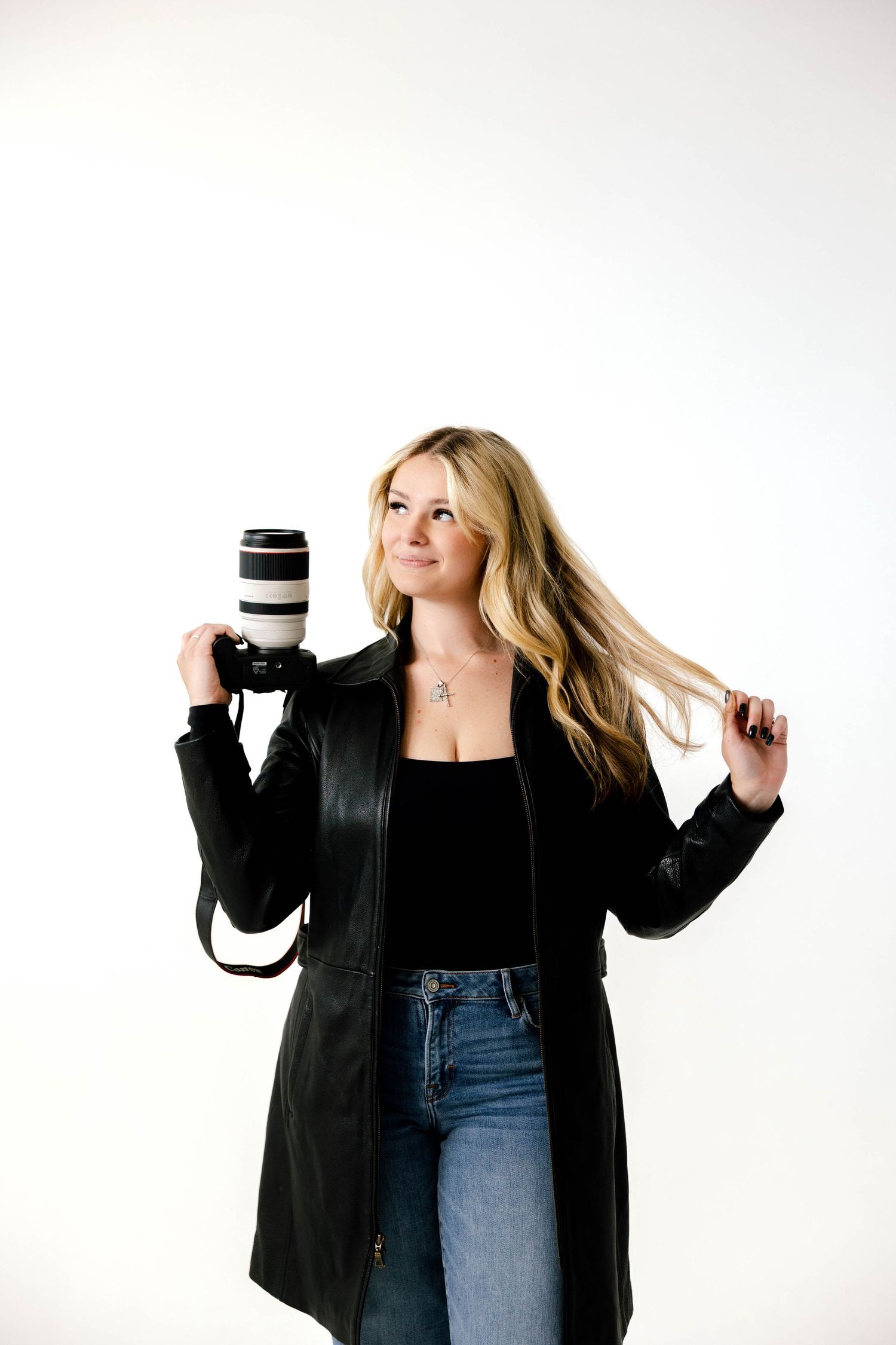 Photographer in black jacket and jeans poses playfully with camera equipment against white background.