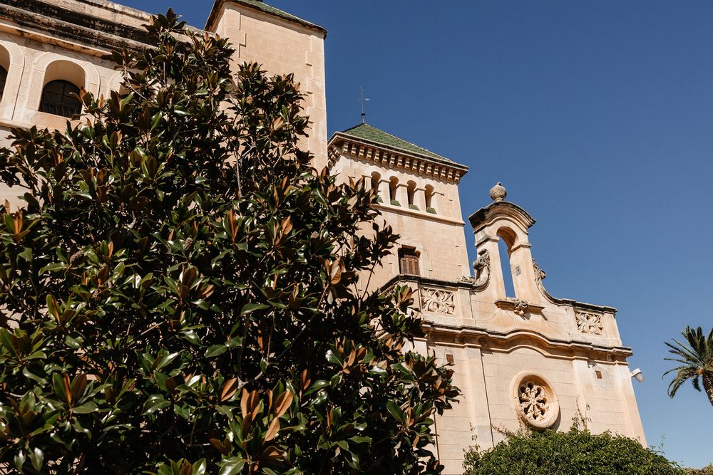 Mediterranean church facade with bell tower and orange trees in the foreground against bright blue sky.