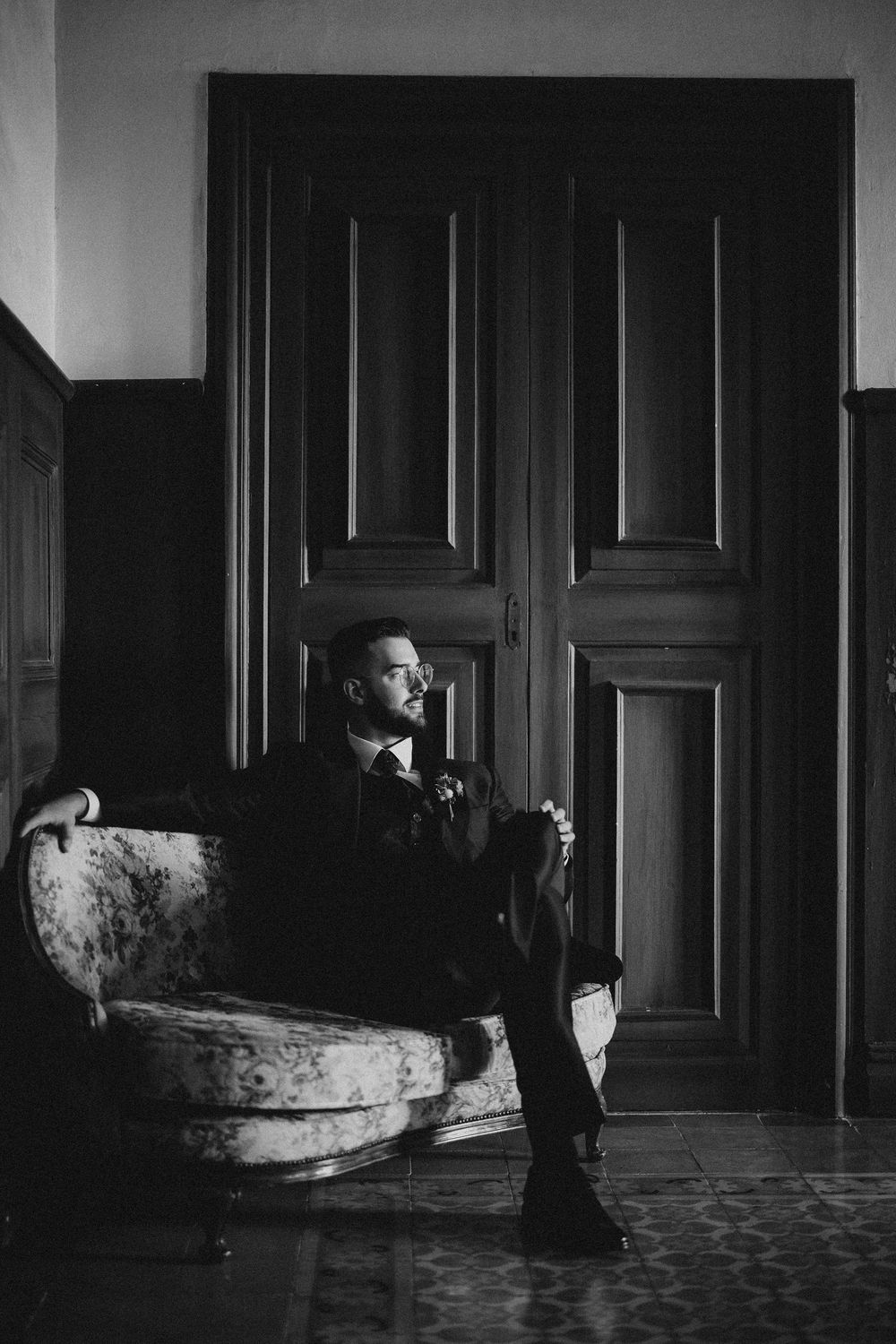 Moody black and white portrait of person sitting in antique armchair against tall wooden doors in dramatic lighting.