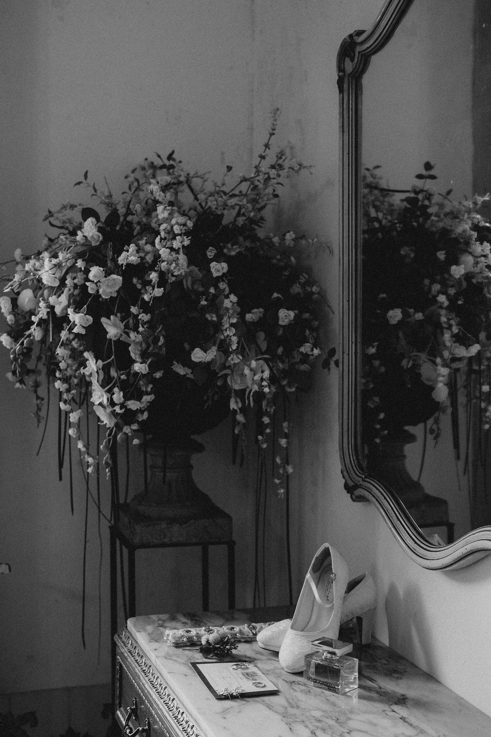 Black and white photo of elegant bridal shoes on a marble table with floral arrangements and ornate mirror decor.