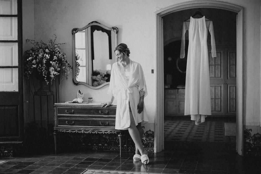 Black and white photograph showing a bridal suite interior with an antique mirror, bench, and white dress hanging in doorway.