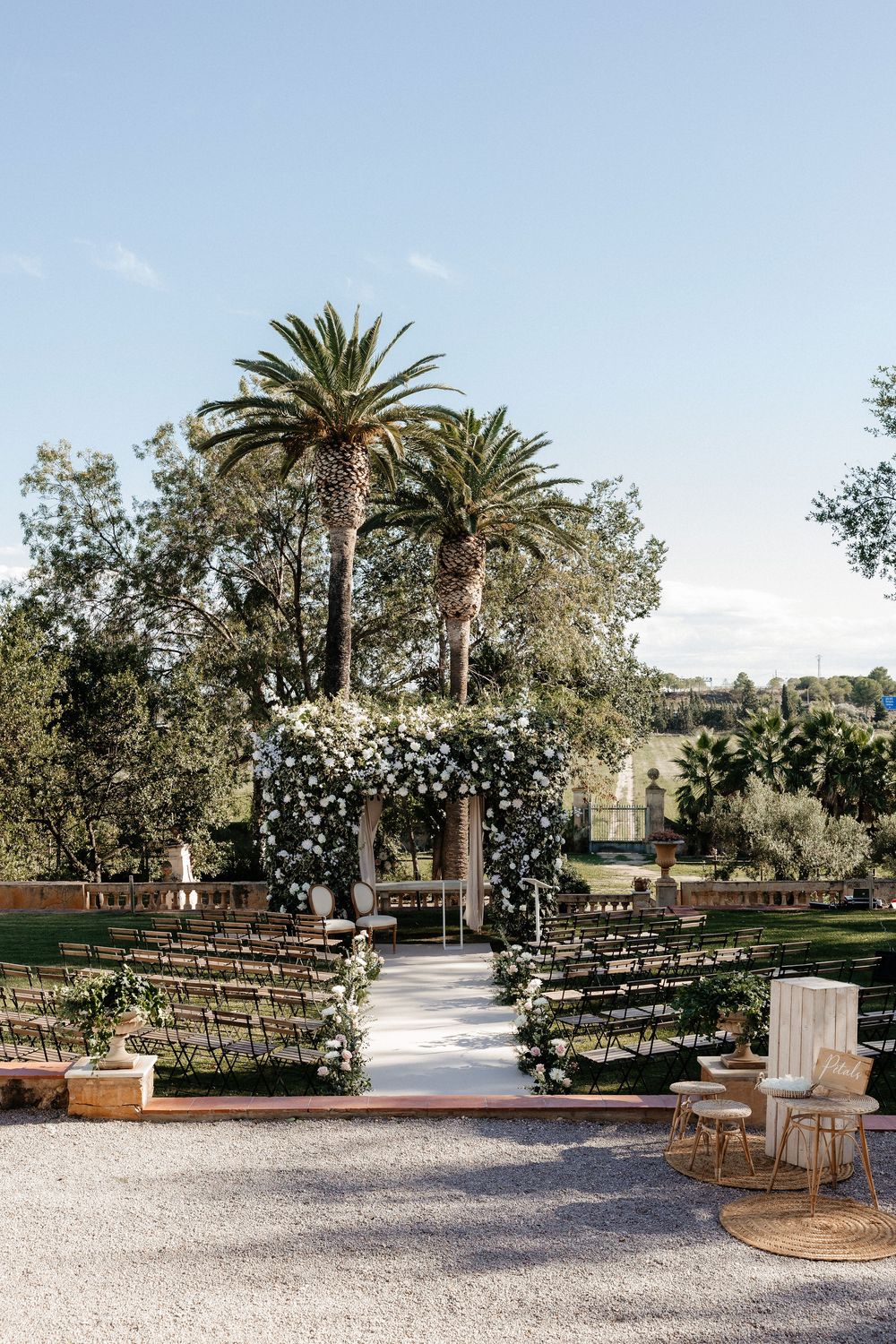 Outdoor wedding ceremony setup with wooden chairs and palm trees in a garden courtyard under natural lighting.